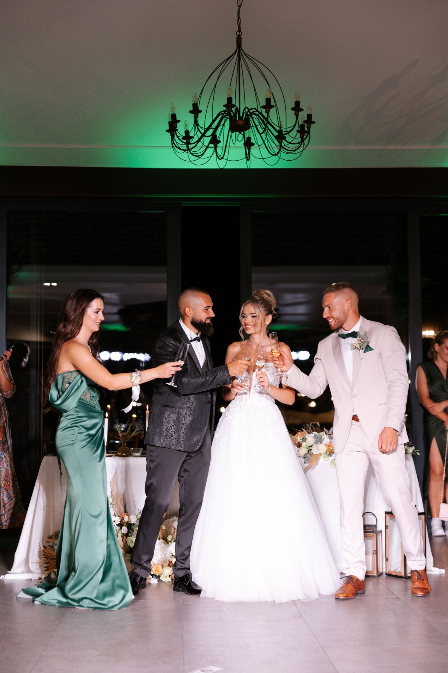 Wedding party toasts and celebrates with drinks under an elegant chandelier with green ambient lighting in the background.