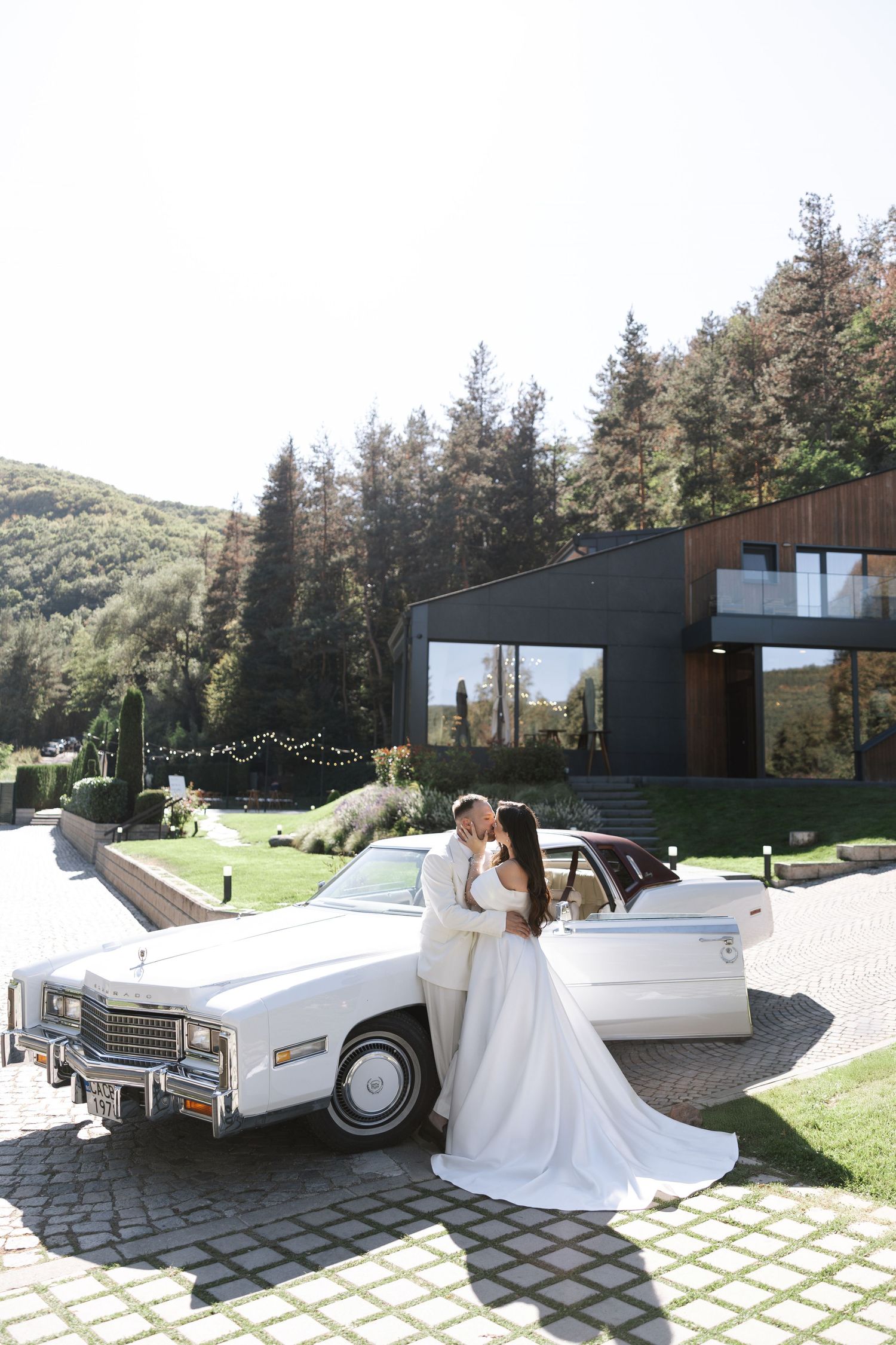 Classic white Cadillac convertible parked in front of modern black building with pine forest backdrop.