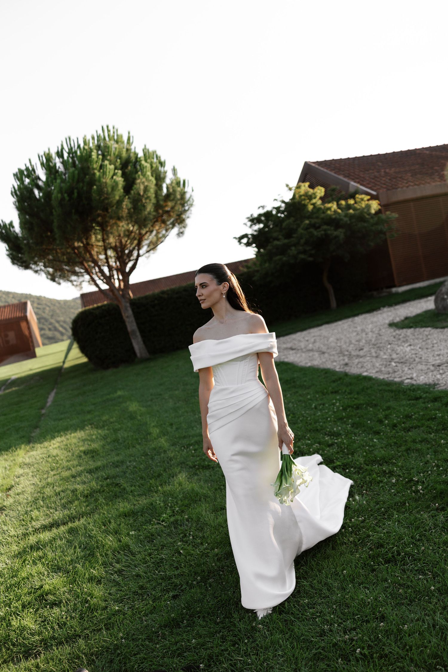 Elegant off-shoulder white wedding dress on a grassy lawn at sunset with a tree in the background.