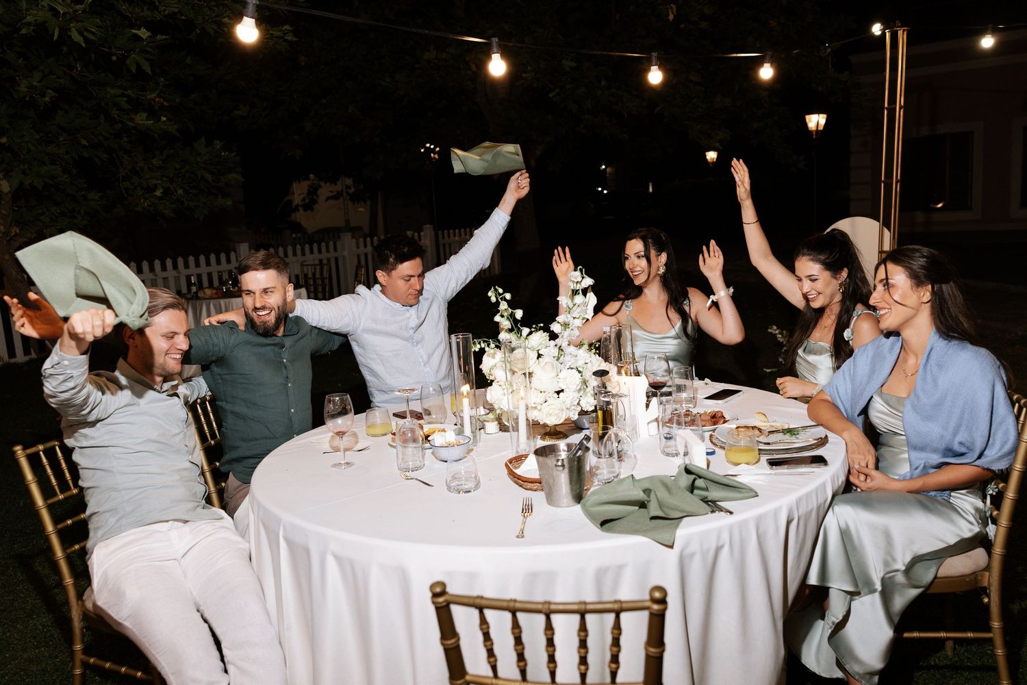 Wedding guests celebrate at an outdoor evening reception with string lights and white tablecloths.