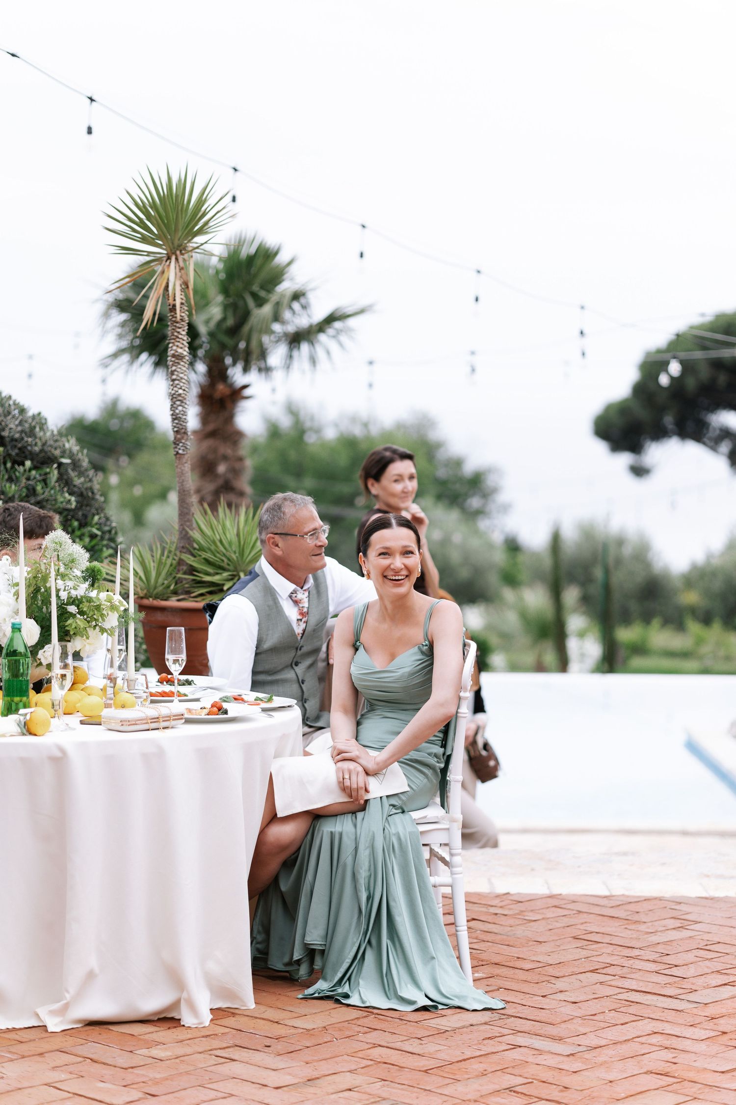 Couple in sage green formal attire share an intimate moment at an outdoor reception with white tables and palm trees.