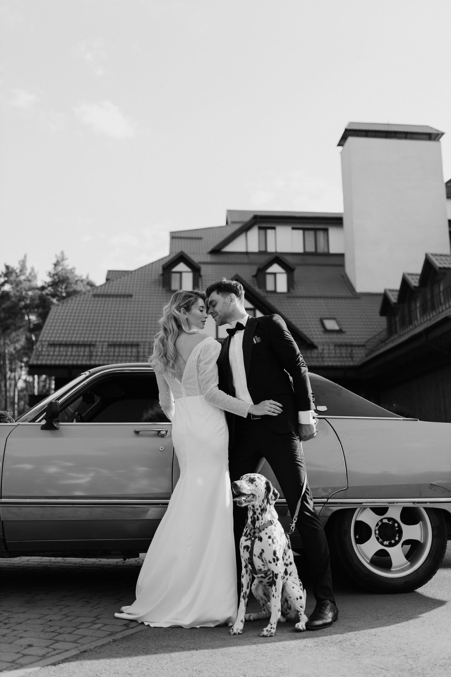 A black and white wedding photo featuring a couple with their Dalmatian dog next to a classic car.