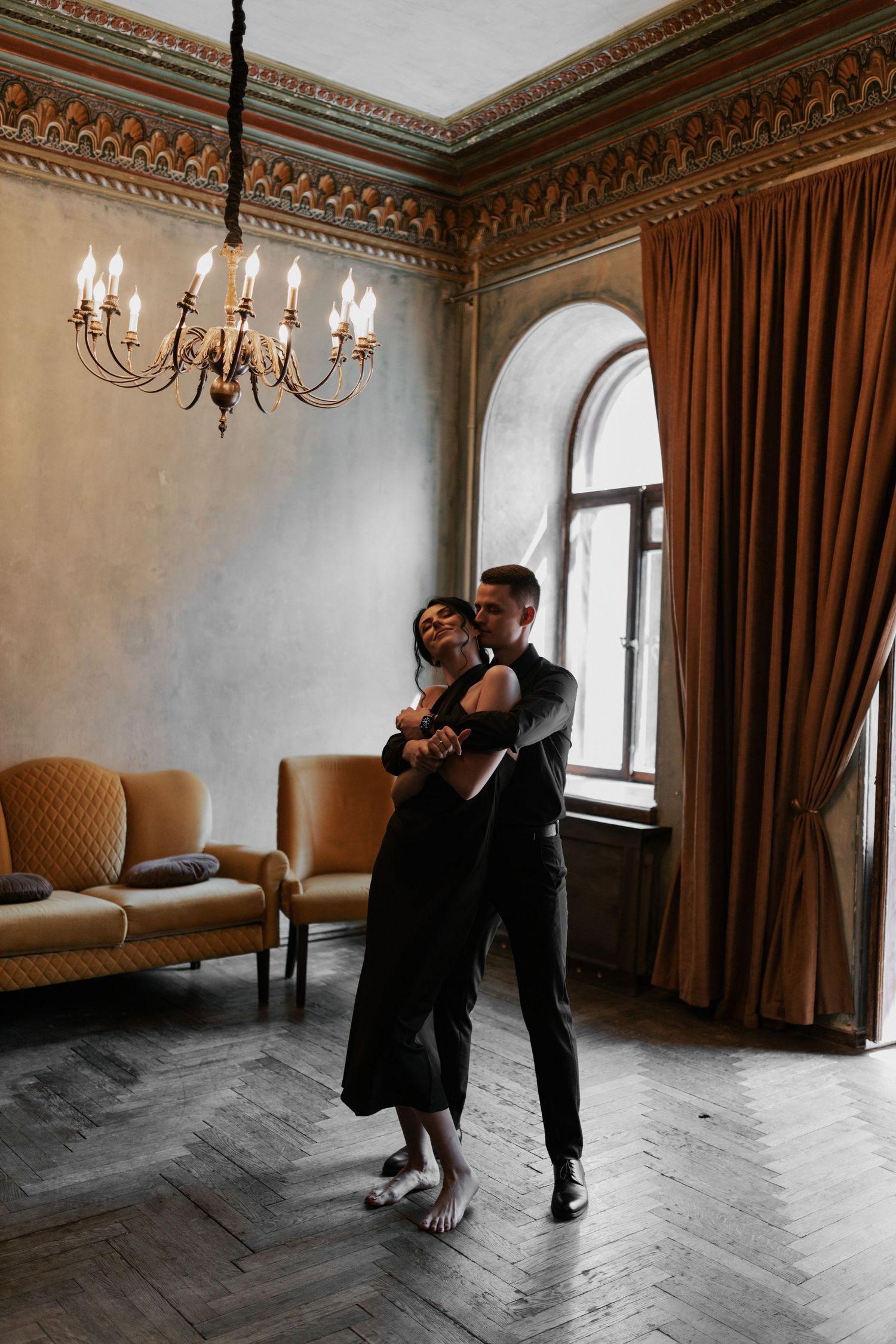 Two dancers embrace in an elegant room with ornate molding, chandelier and herringbone floors.