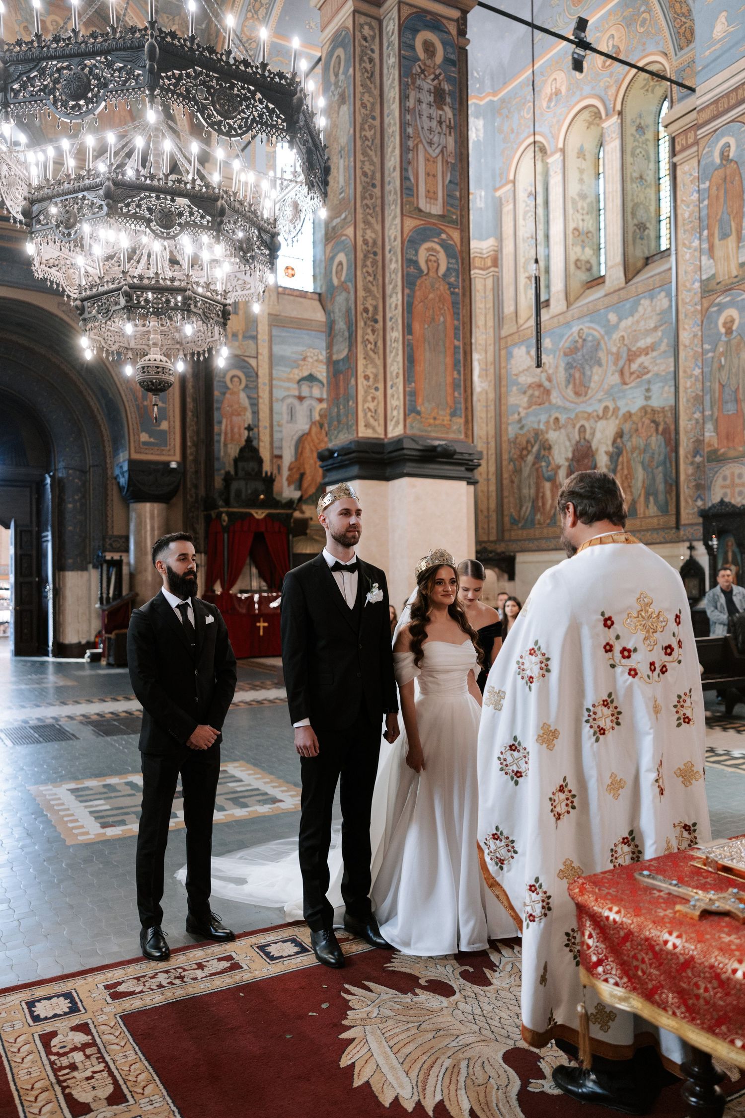 Orthodox wedding ceremony in progress beneath grand chandeliers and painted church walls.