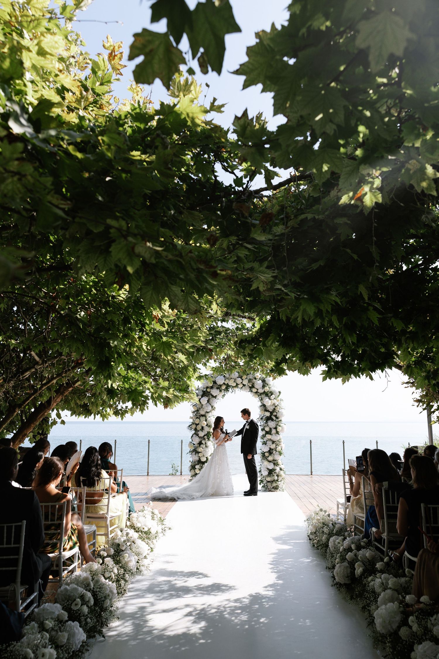 A wedding ceremony setup under a shaded canopy with white floral arrangements and ocean views in the background.