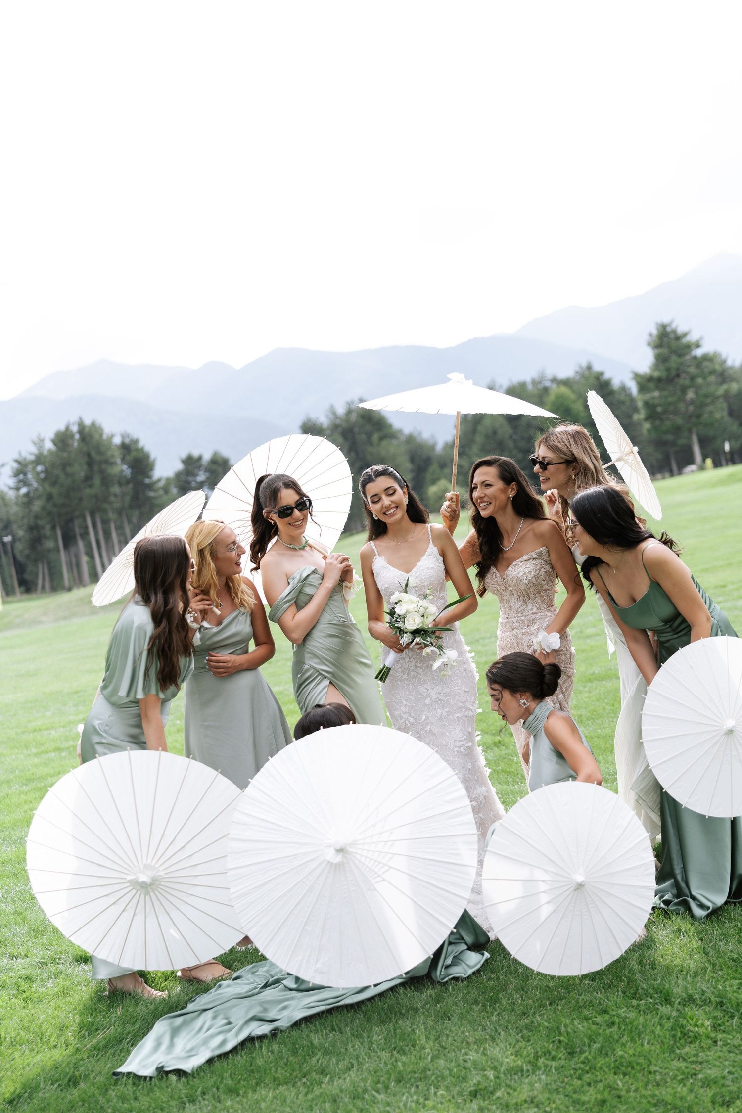 Group poses with white paper parasols on a grassy lawn with mountains in the background during a summer celebration.