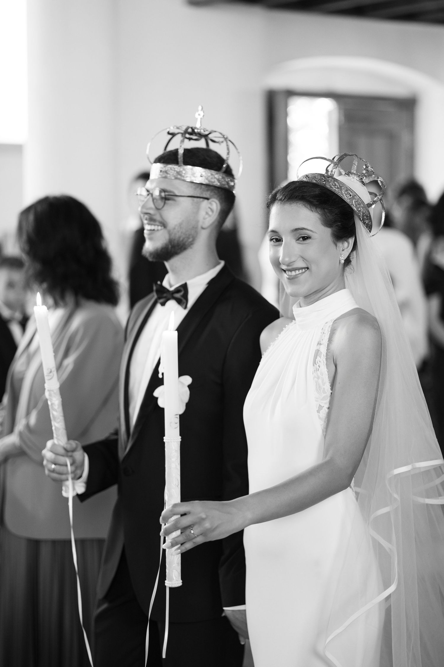A black and white wedding ceremony photo showing a bride and groom wearing crowns and holding candles during an Orthodox service.