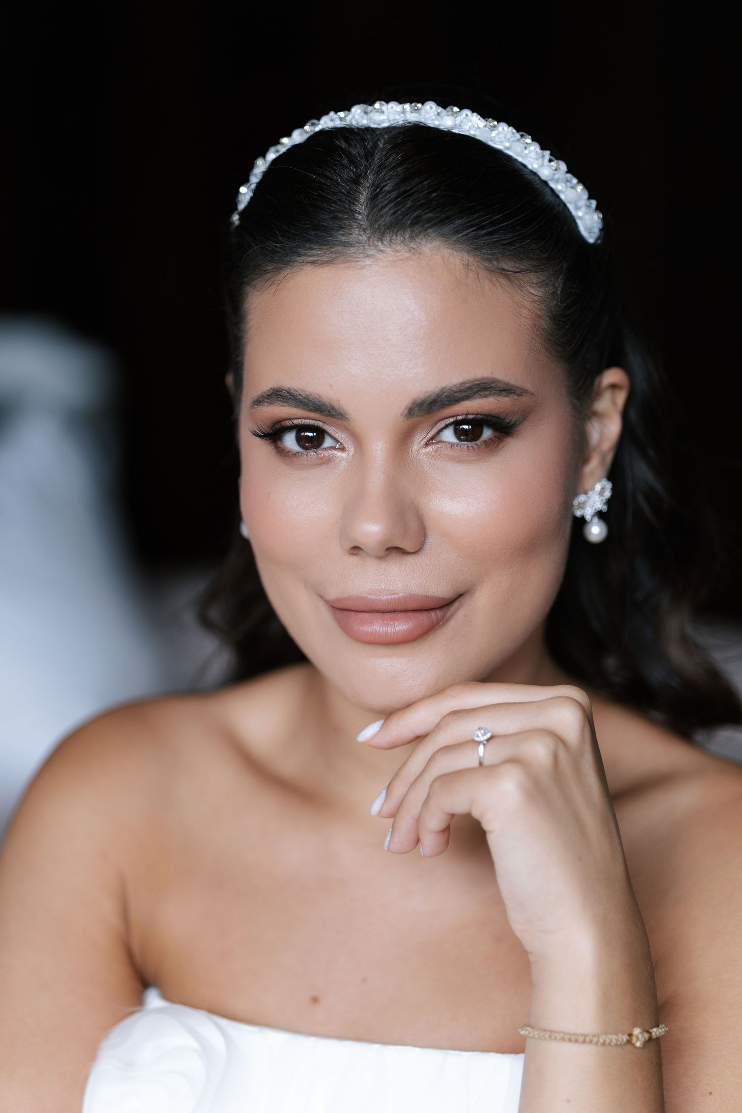 A close-up portrait of someone wearing bridal makeup and jewelry against a dark background.
