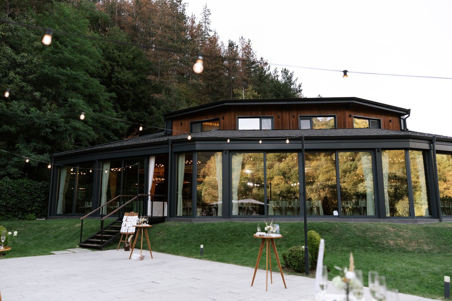 Modern glass-walled event venue with floor-to-ceiling windows nestled against a forest backdrop at dusk.