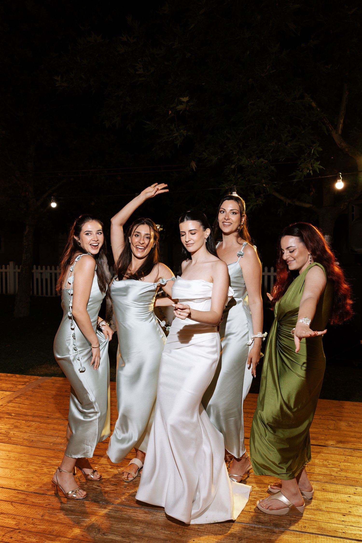 A group of people in white dresses dancing together on a wooden outdoor dance floor at night.