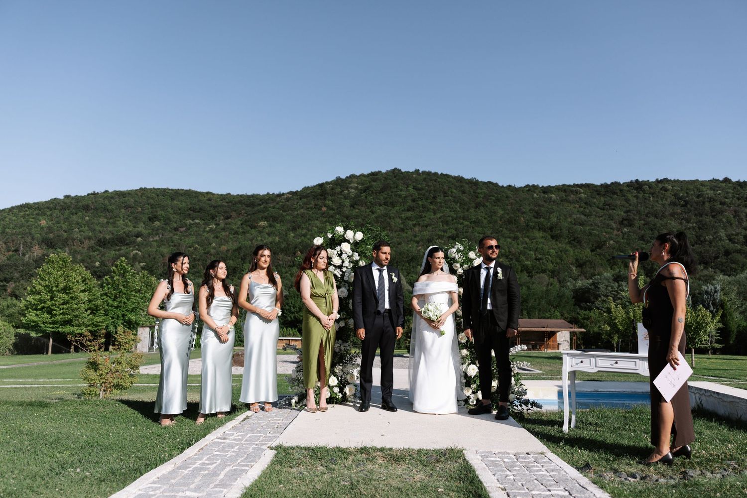 Wedding party stands on an outdoor ceremony aisle with green mountains and trees in the background.