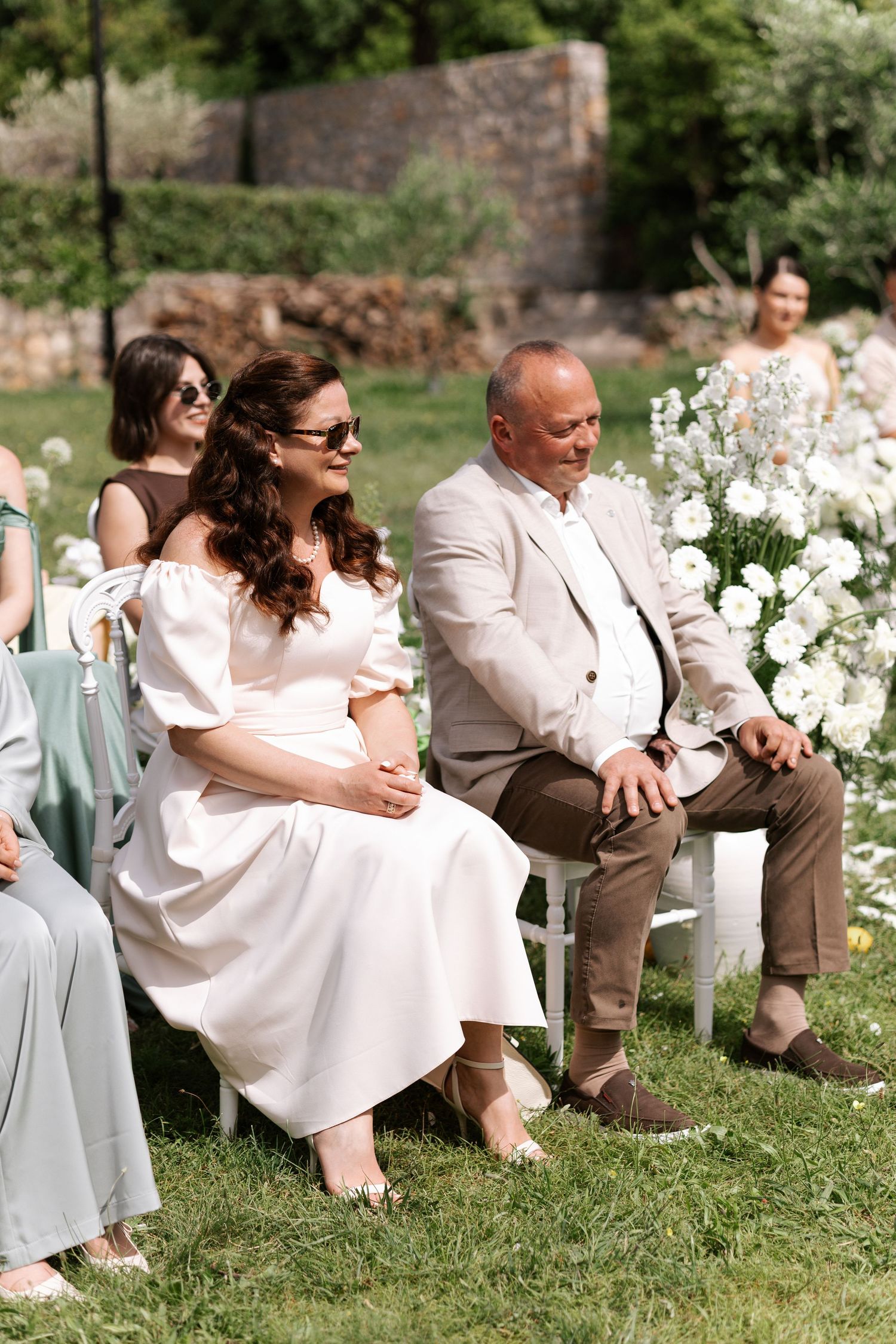 Wedding guests seated outdoors on white chairs near floral arrangements during a garden ceremony.