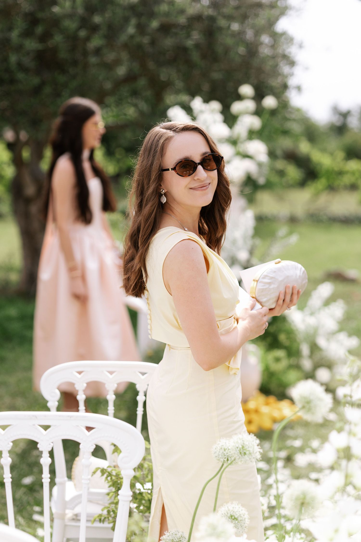 Woman in yellow dress poses in garden with white flowers and greenery on sunny summer day.