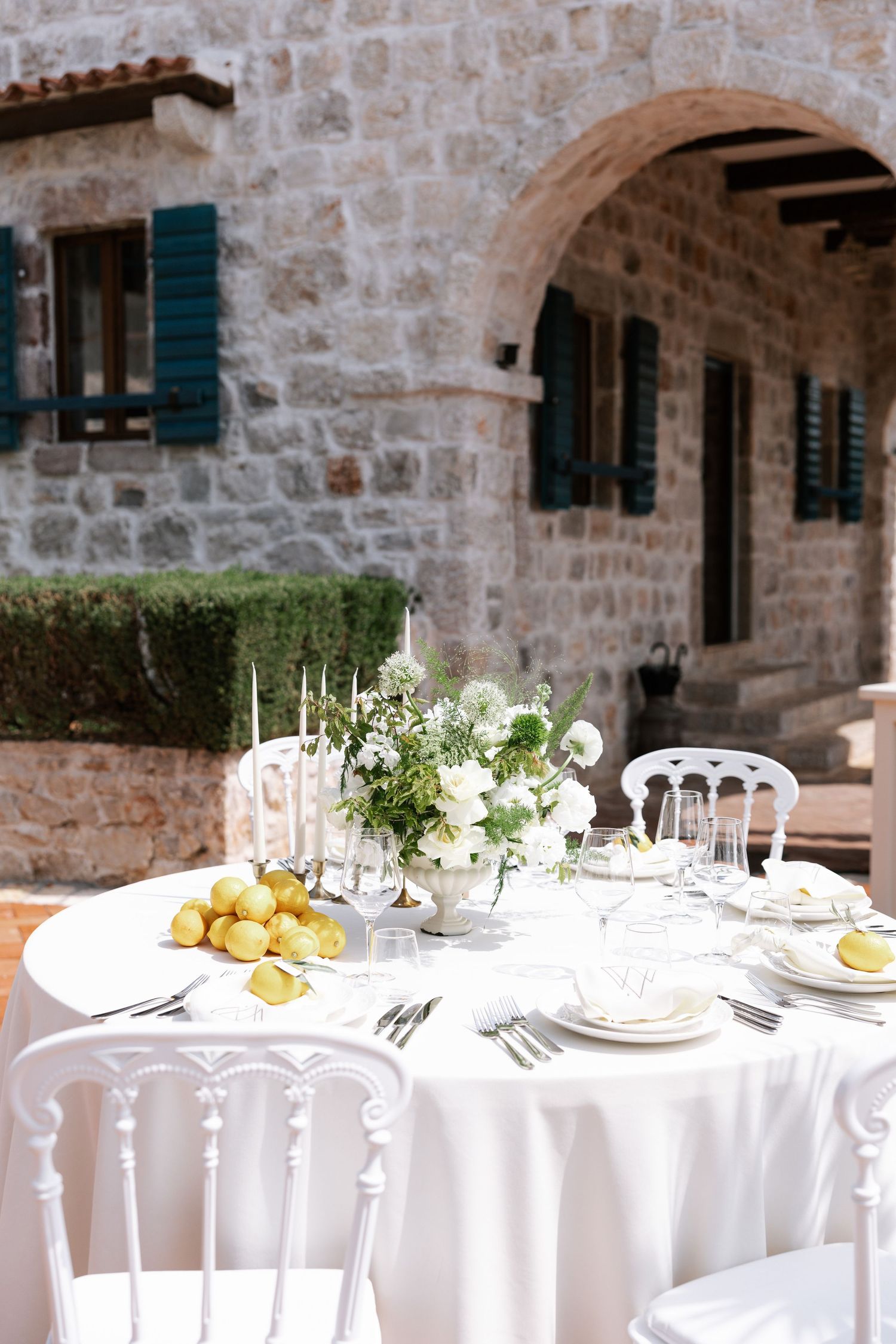 Elegant outdoor table setting with white linens, candles and lemons at a Mediterranean stone villa with blue shutters.