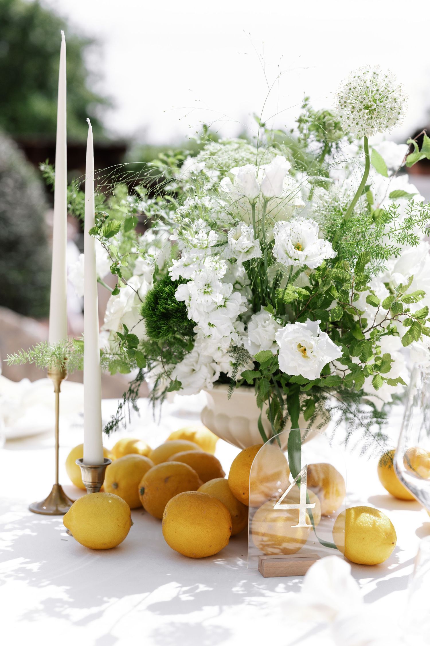 Elegant table setting with white floral centerpiece, tall white candles, and fresh lemons scattered on white tablecloth.