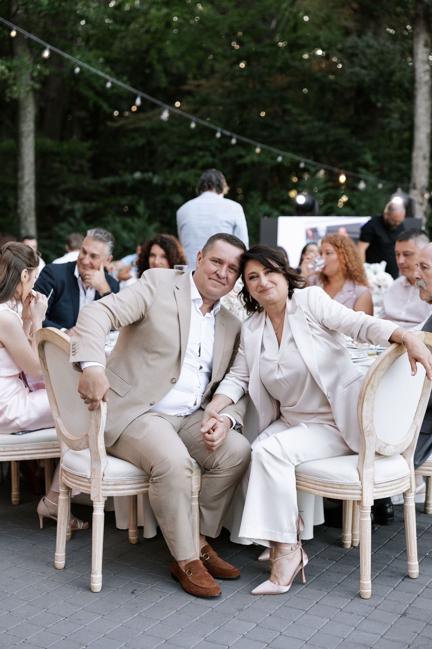 Wedding party in cream-colored attire posing playfully on chairs outdoors.
