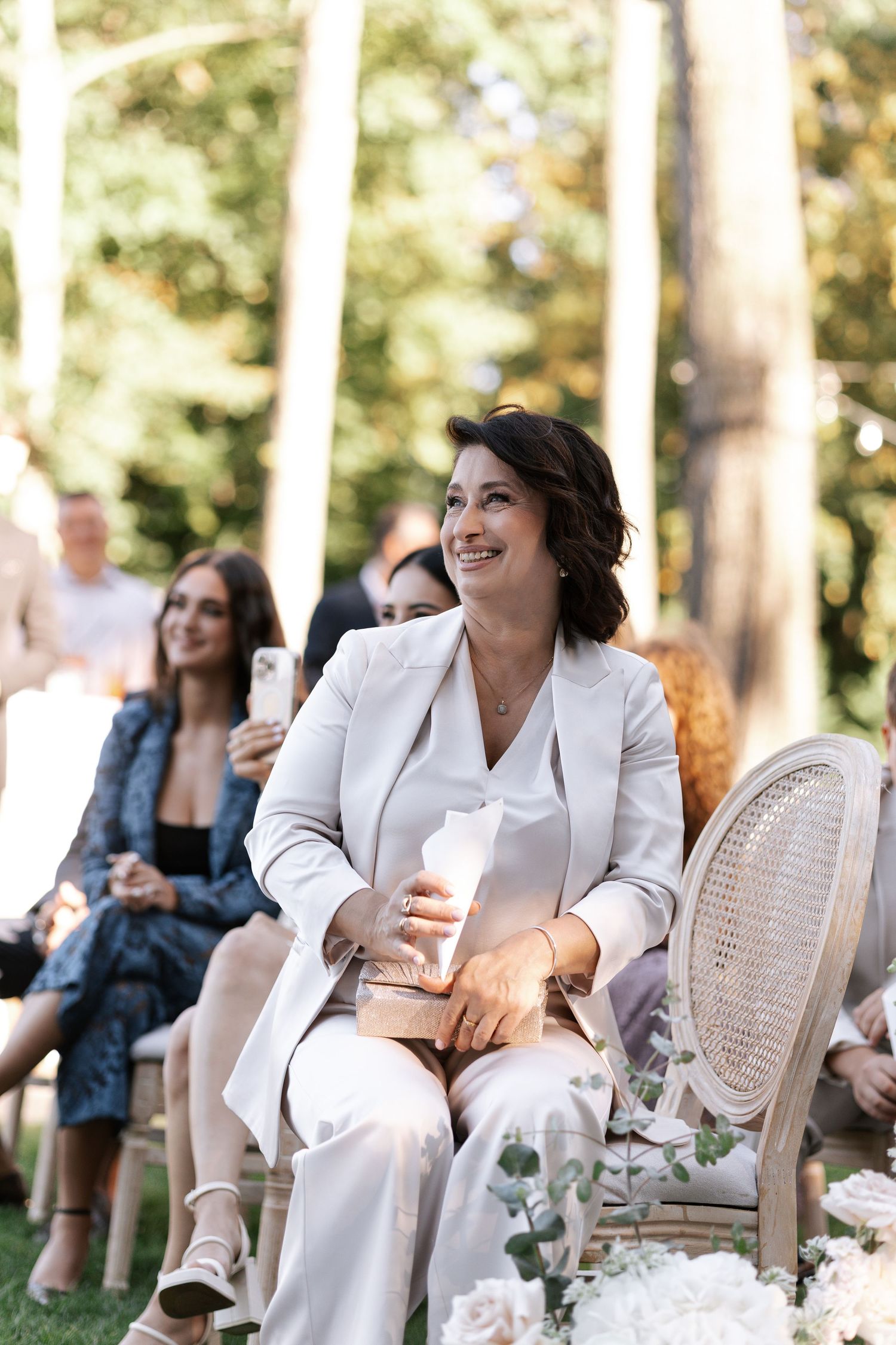 Outdoor wedding reception seating with elegant white chairs and natural lighting through trees.