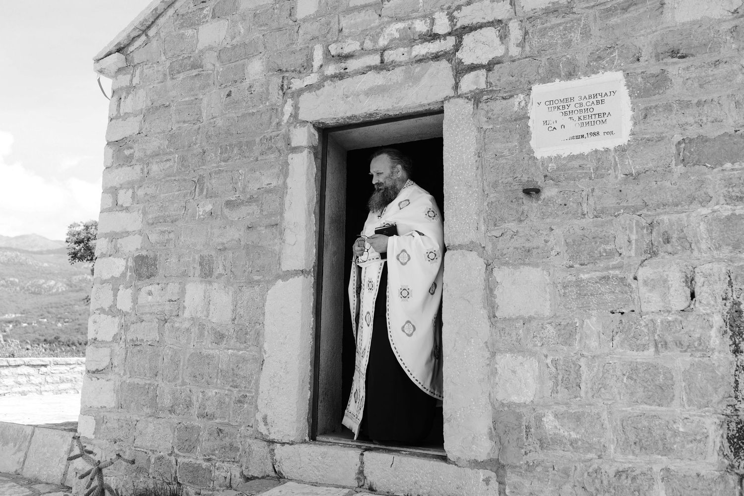 A black and white photograph shows three people looking out of windows in an old stone building wall.