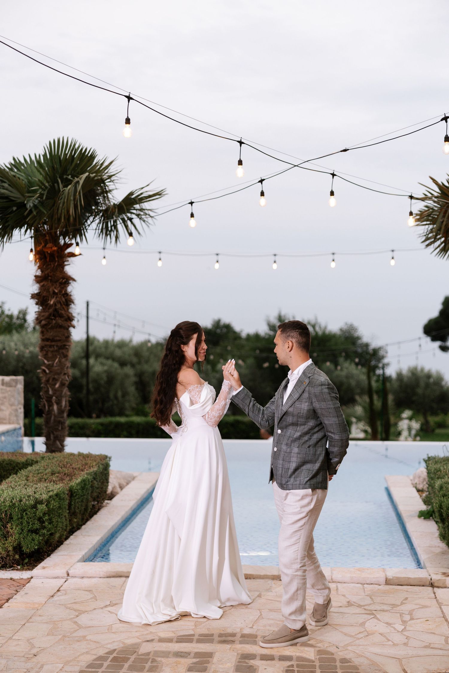 A romantic outdoor wedding moment captured on a stone patio with string lights and palm trees by a pool.