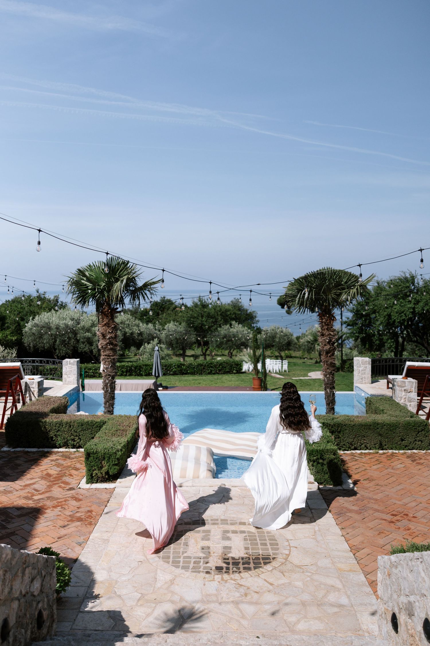 Two women in flowing dresses stand on a stone pathway overlooking a pool with palm trees and string lights at an outdoor venue.