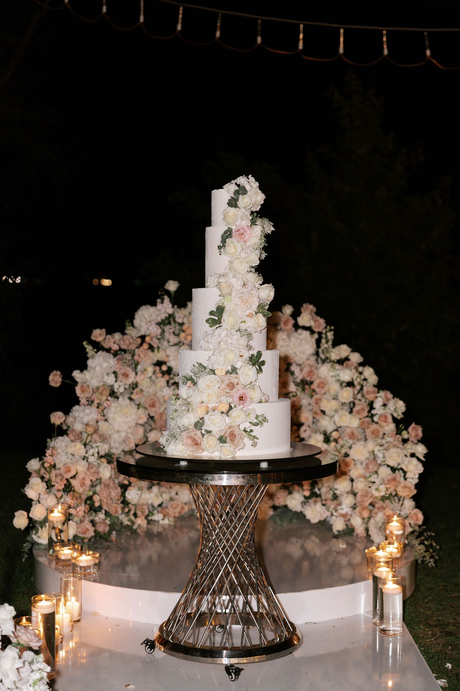 An elegant tiered white wedding cake decorated with cascading pink and peach roses sits on a metallic geometric stand.