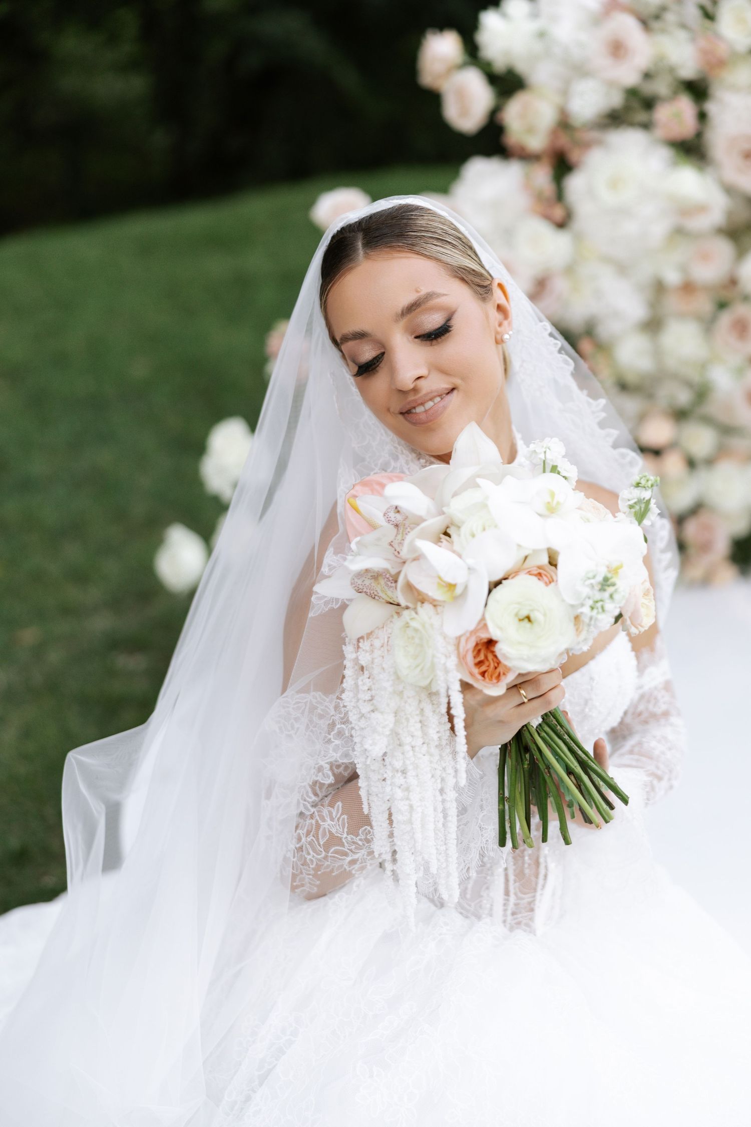 Bride in white lace gown holds romantic peach and white bouquet against floral backdrop.