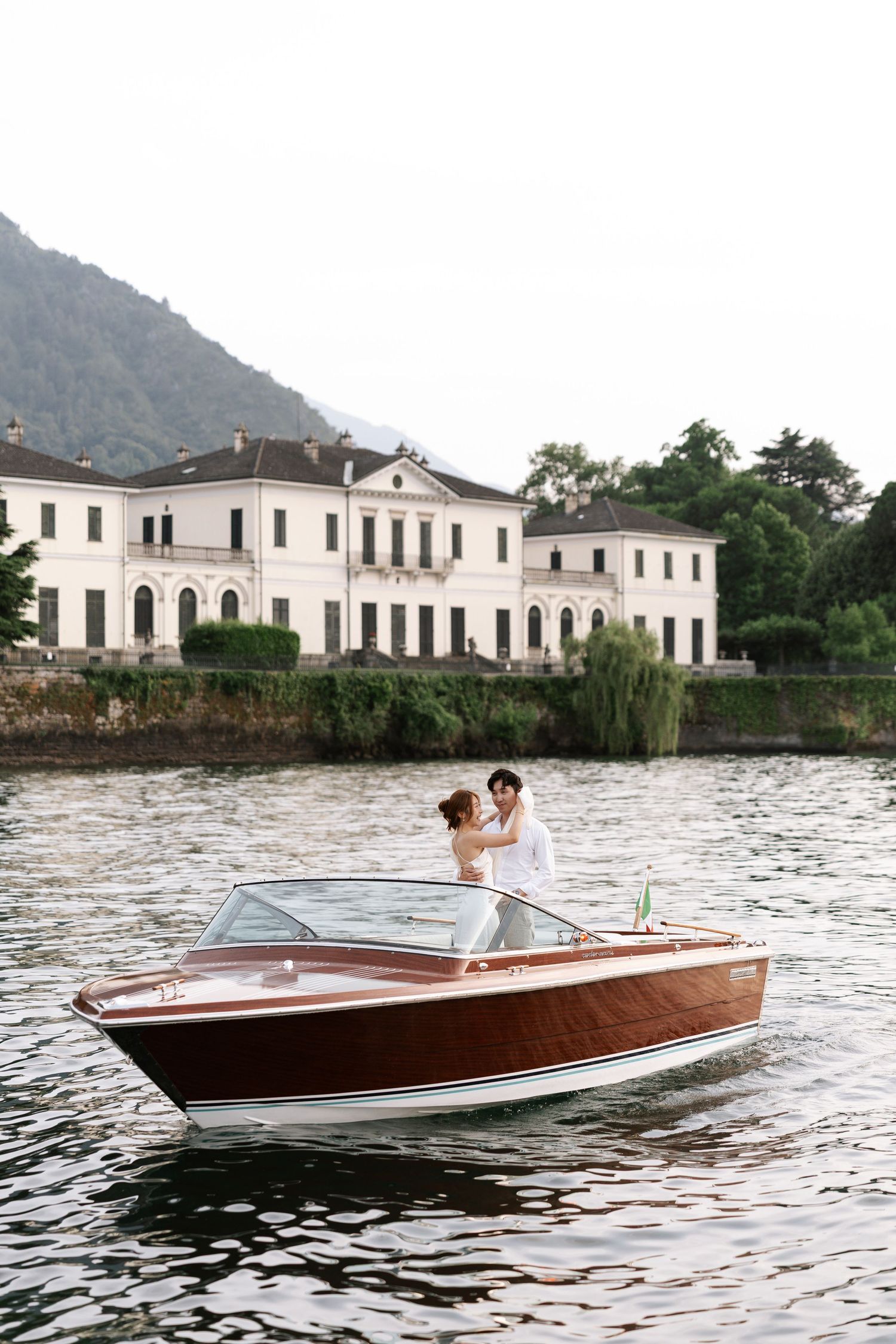 Newlyweds enjoy a boat ride in a classic wooden speedboat on Lake Como with historic villa in background.