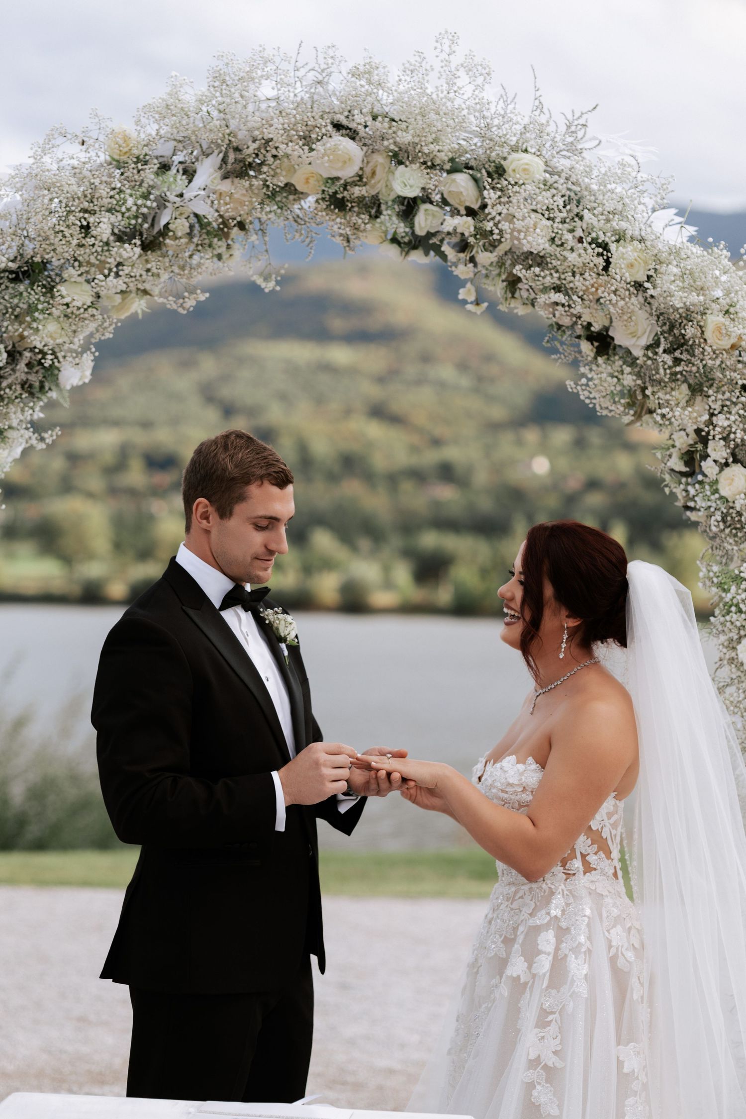 Couple exchanges rings under an elegant white floral arch with mountains in the background.