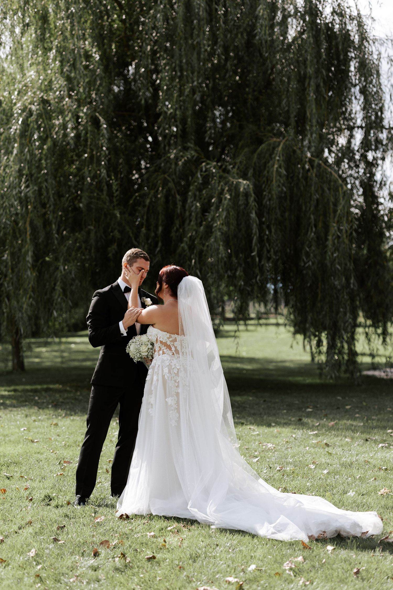 A tender moment between newlyweds on their wedding day beneath a weeping willow tree.