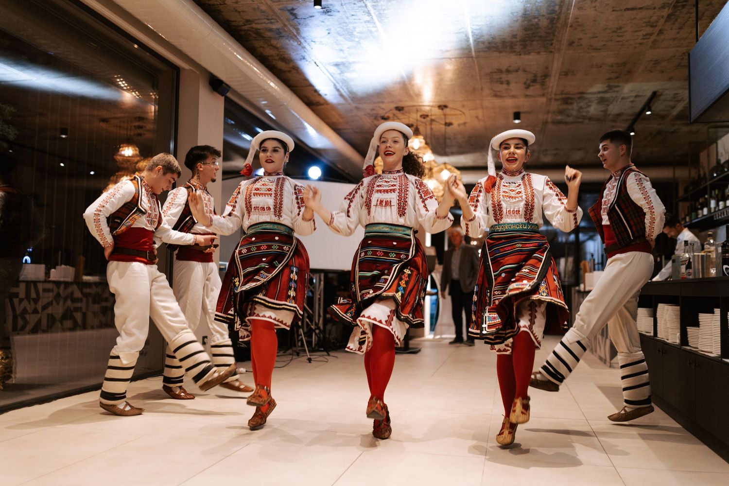 Traditional folk dancers performing in colorful costumes and embroidered vests at an indoor venue.