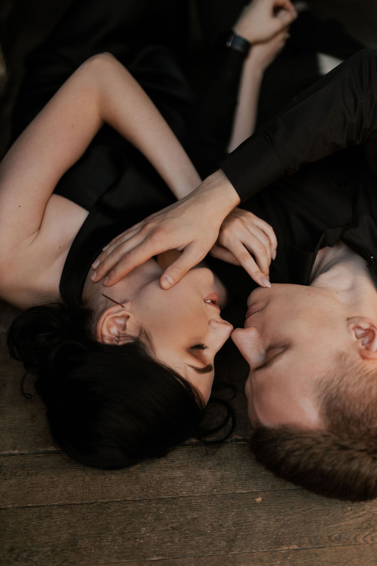 A romantic couple lies on wooden floor in intimate black attire.