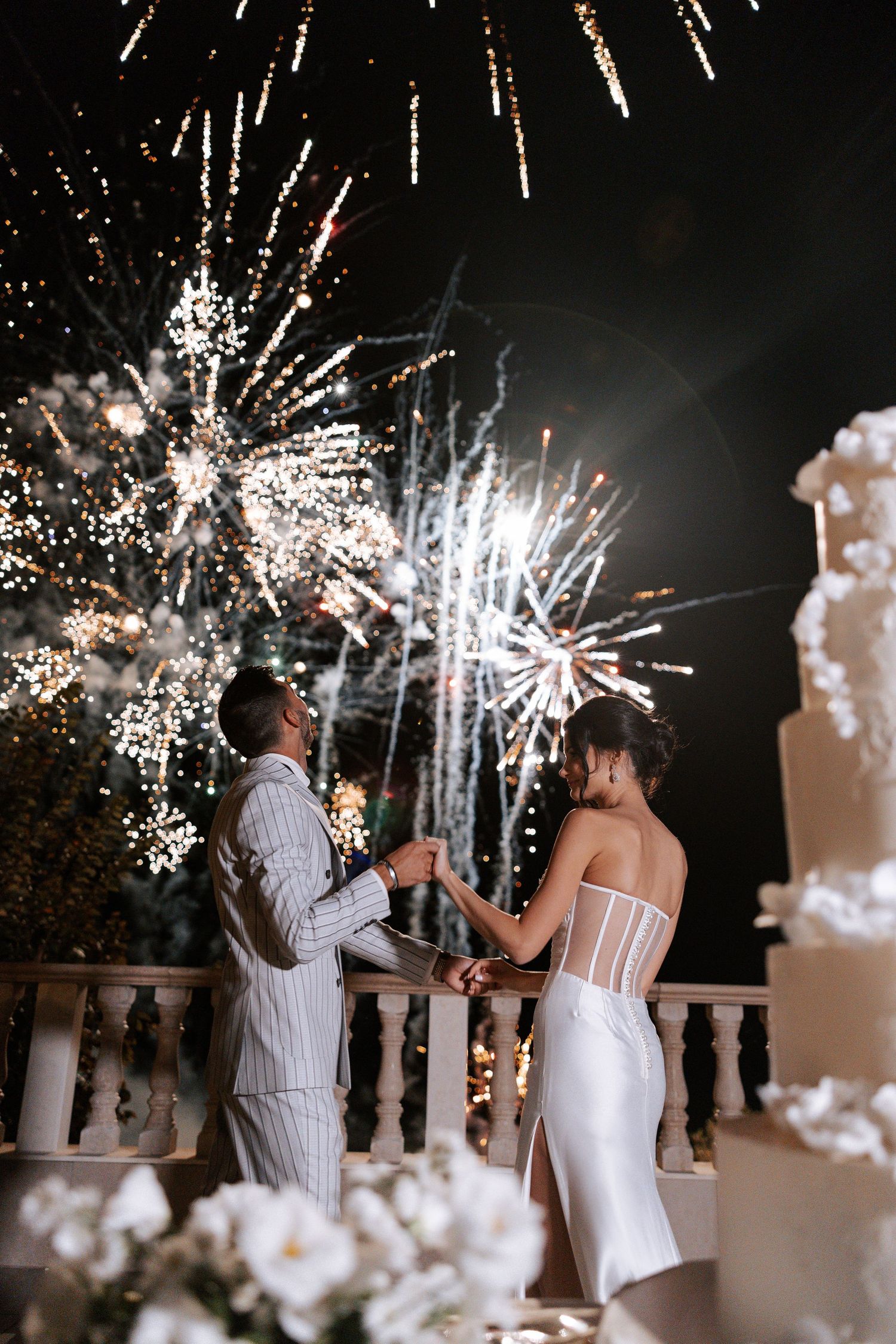 Couple celebrates with spectacular fireworks display and wedding cake during nighttime outdoor reception.