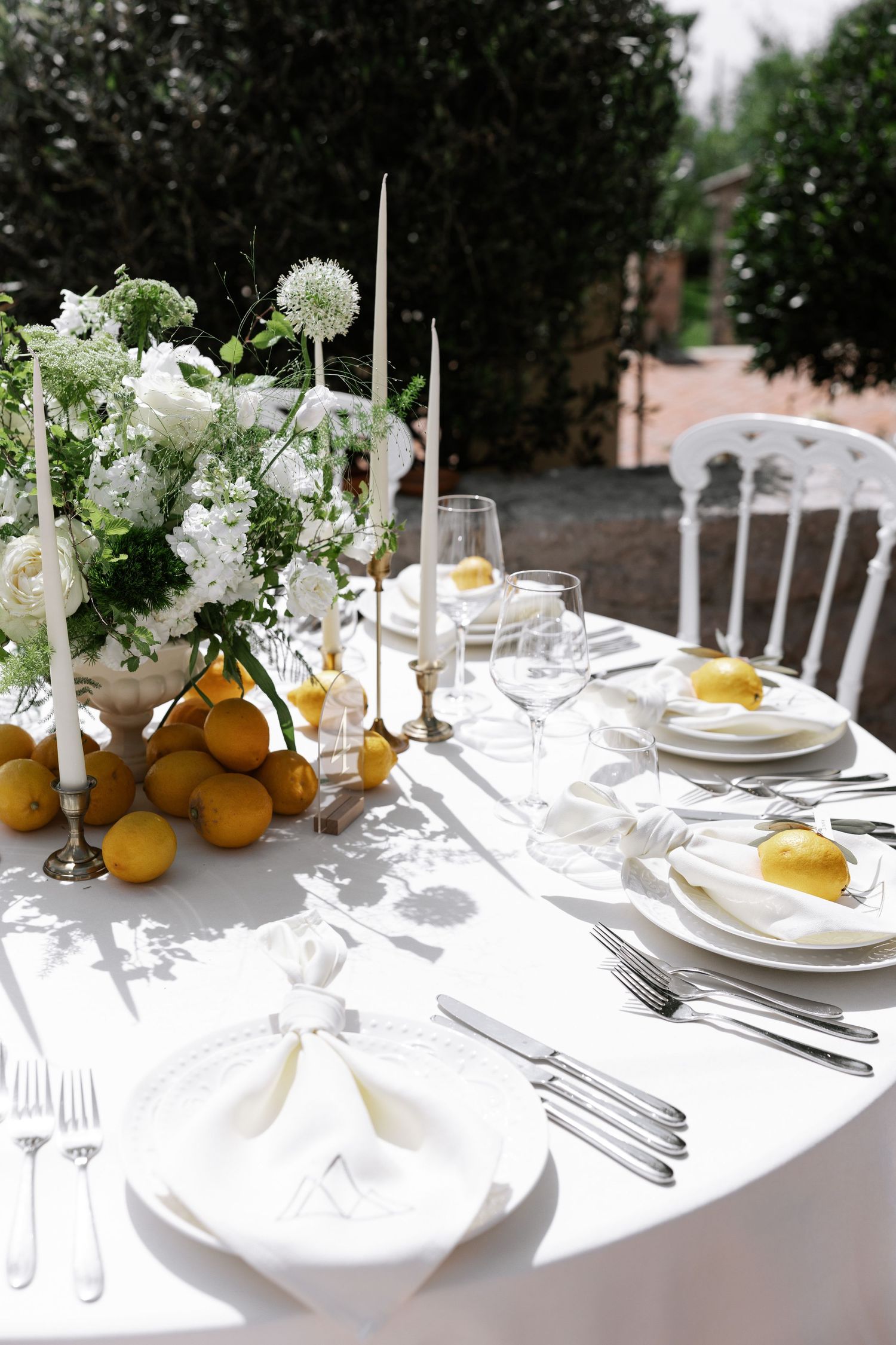 Elegant white table setting with fresh citrus fruits and floral arrangements creating a bright outdoor dining atmosphere.