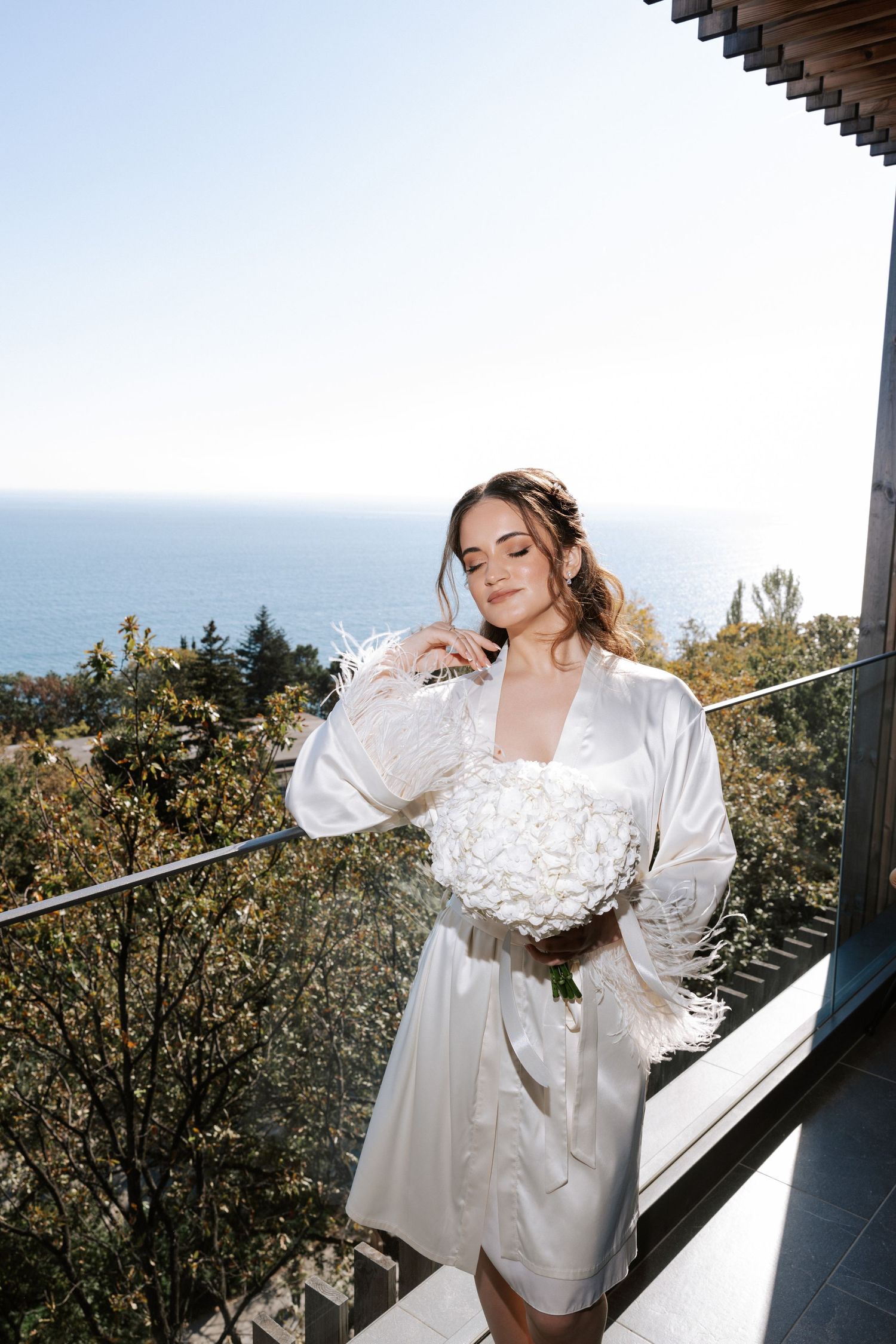 Figure in white silk robe holding white bouquet on oceanview balcony.