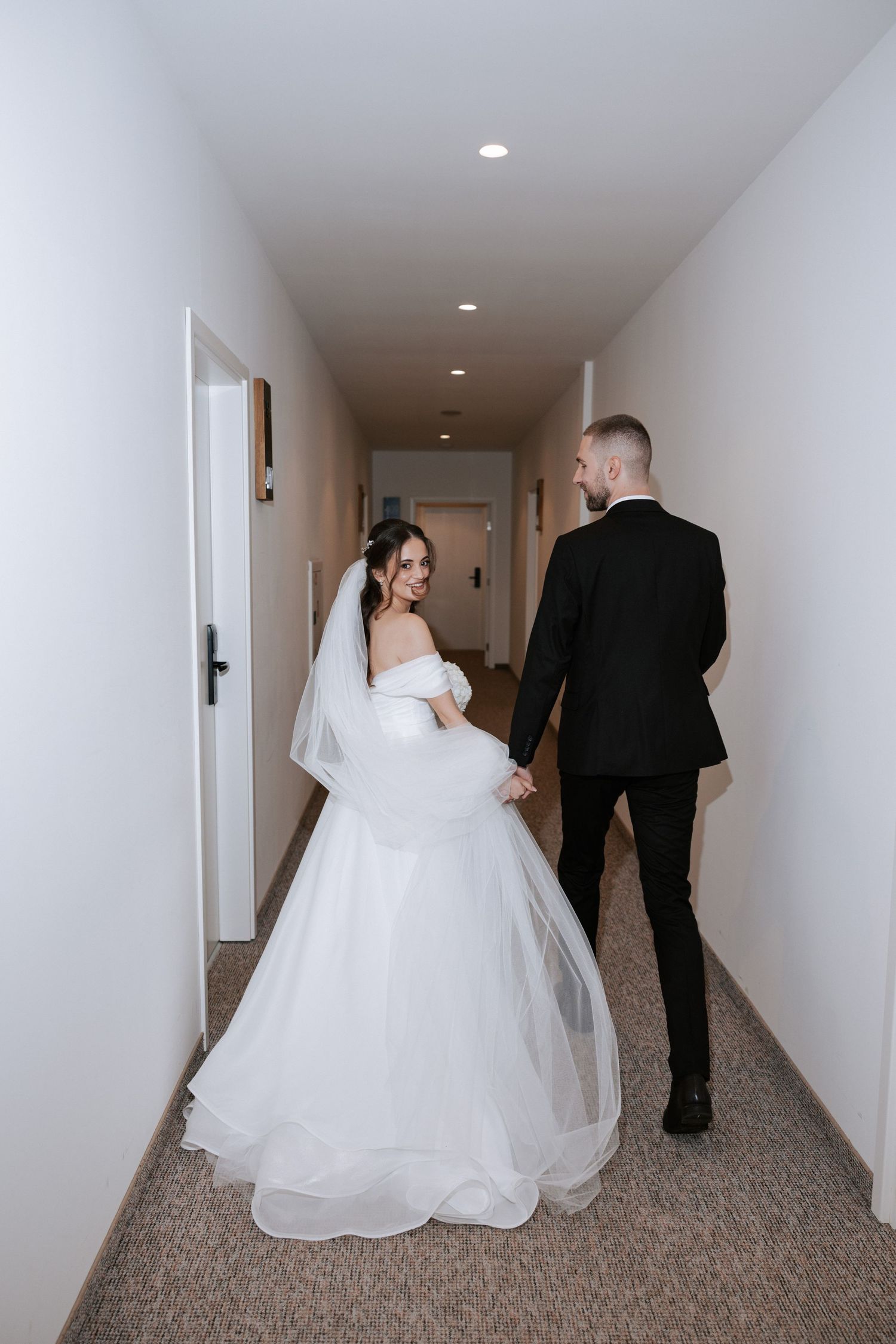 Wedding couple walks hand in hand down a bright hallway with white walls.