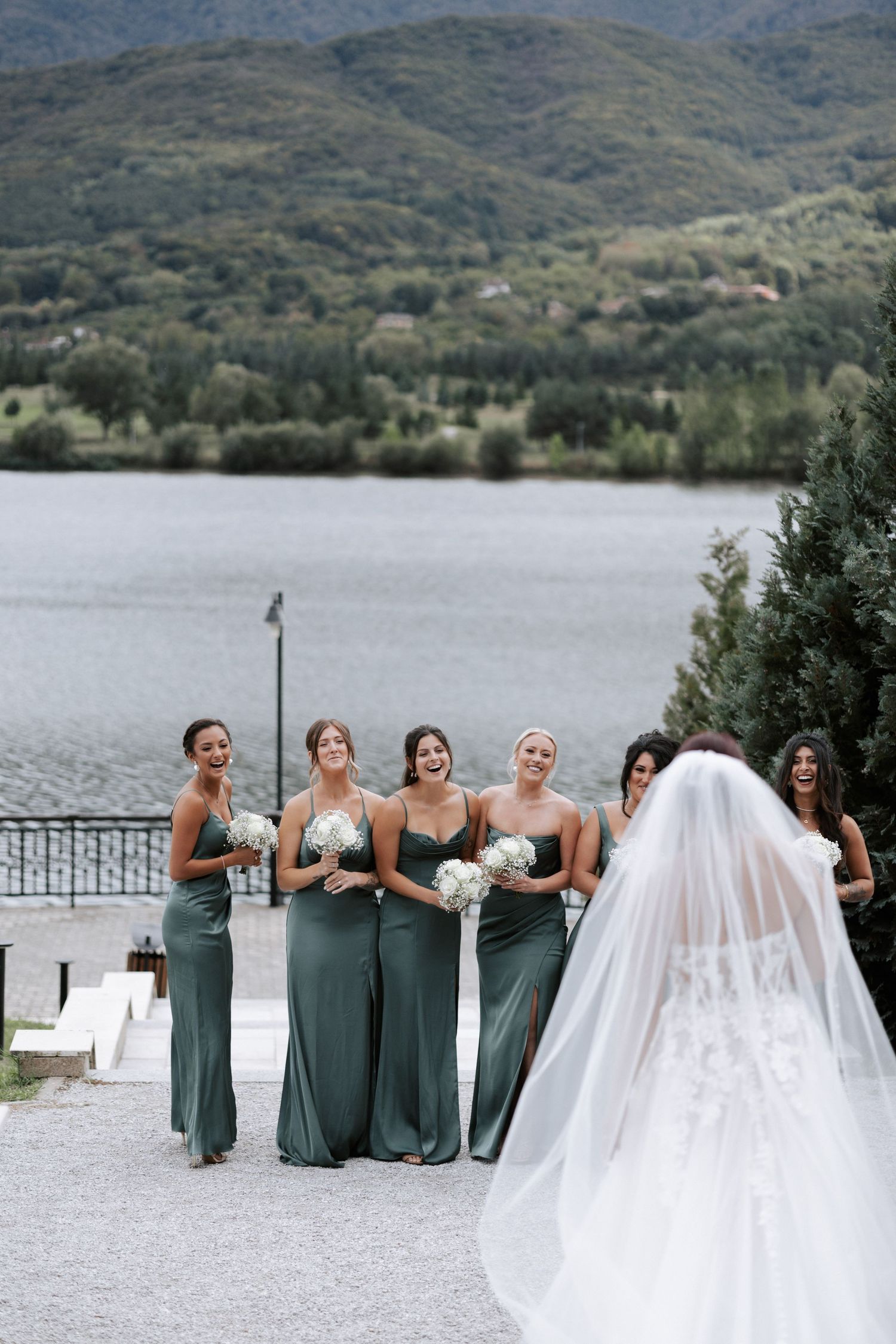 Bridesmaids in sage dresses share a moment with the bride against mountain scenery.