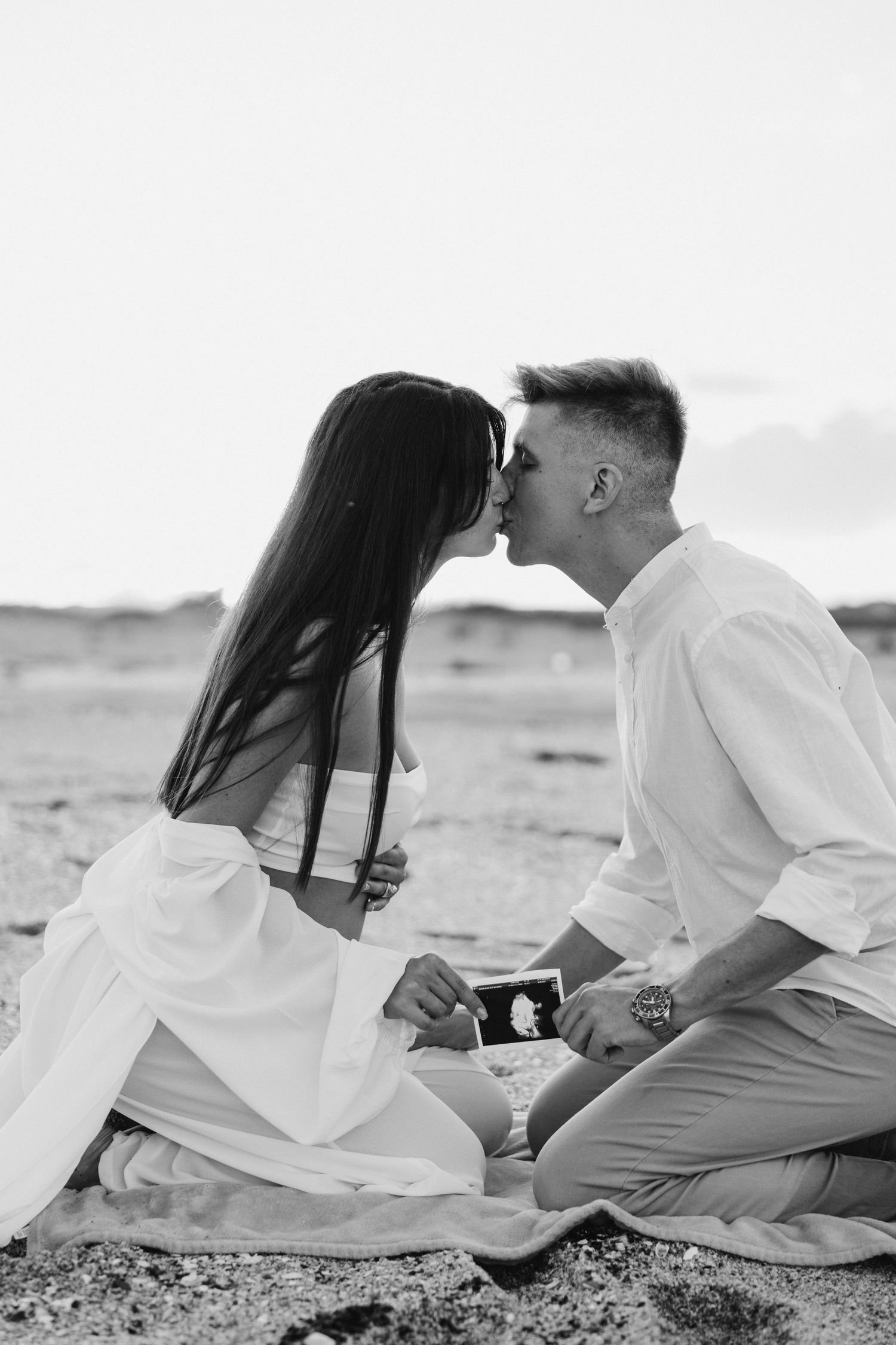 A romantic black and white photo of a couple sharing a kiss on the beach while holding a sonogram.