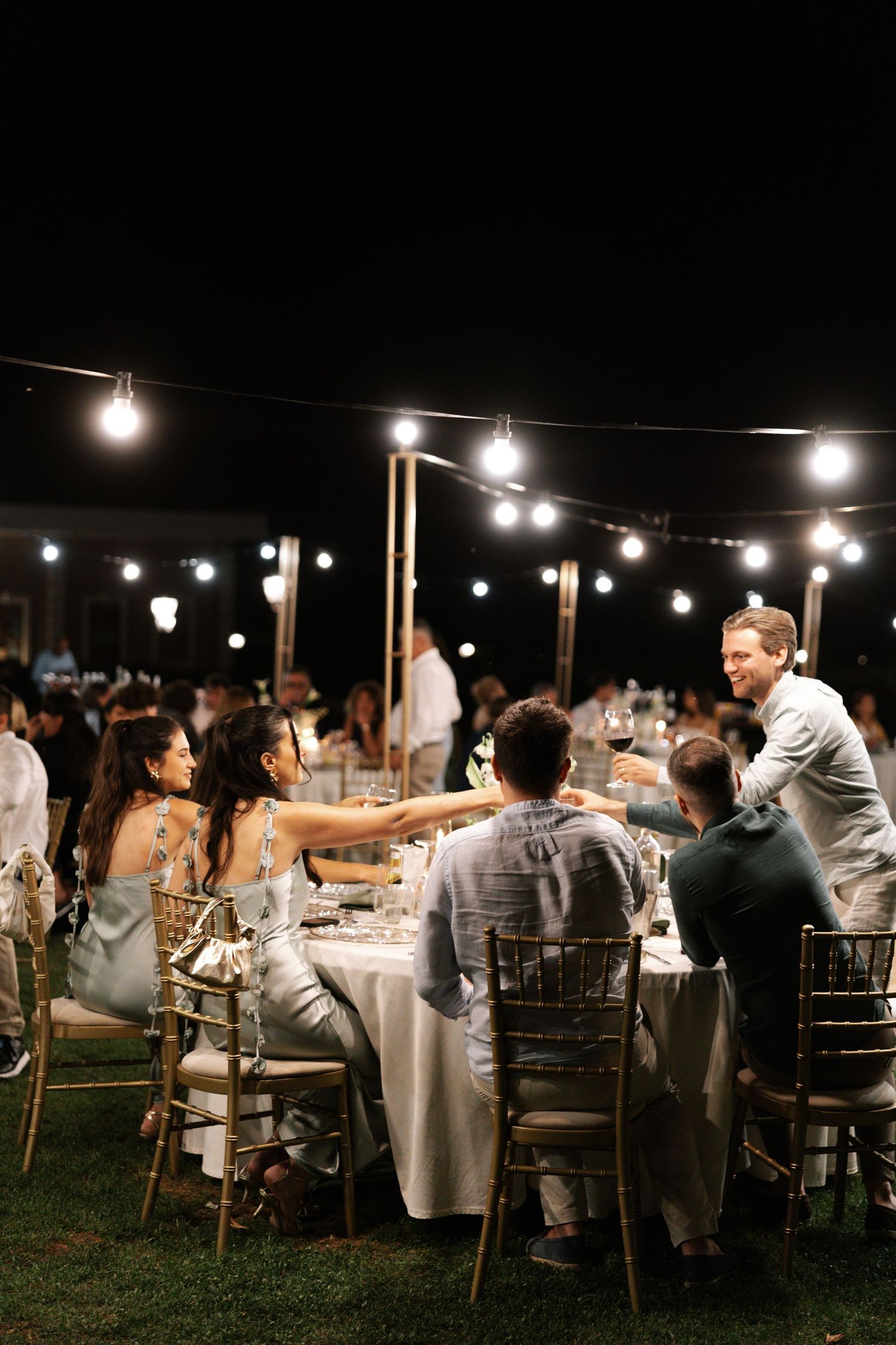 String lights illuminate an outdoor evening wedding reception with guests dining at elegant tables on a lawn.