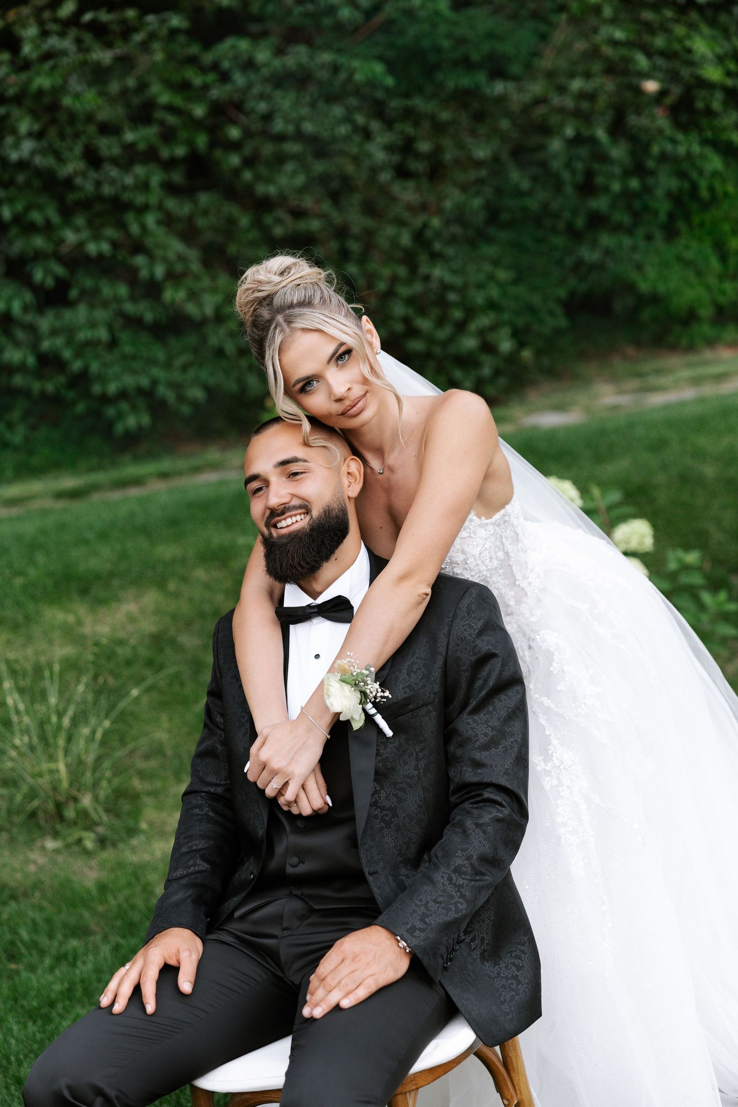 Romantic wedding couple poses playfully outdoors in a garden setting with lush green foliage in the background.