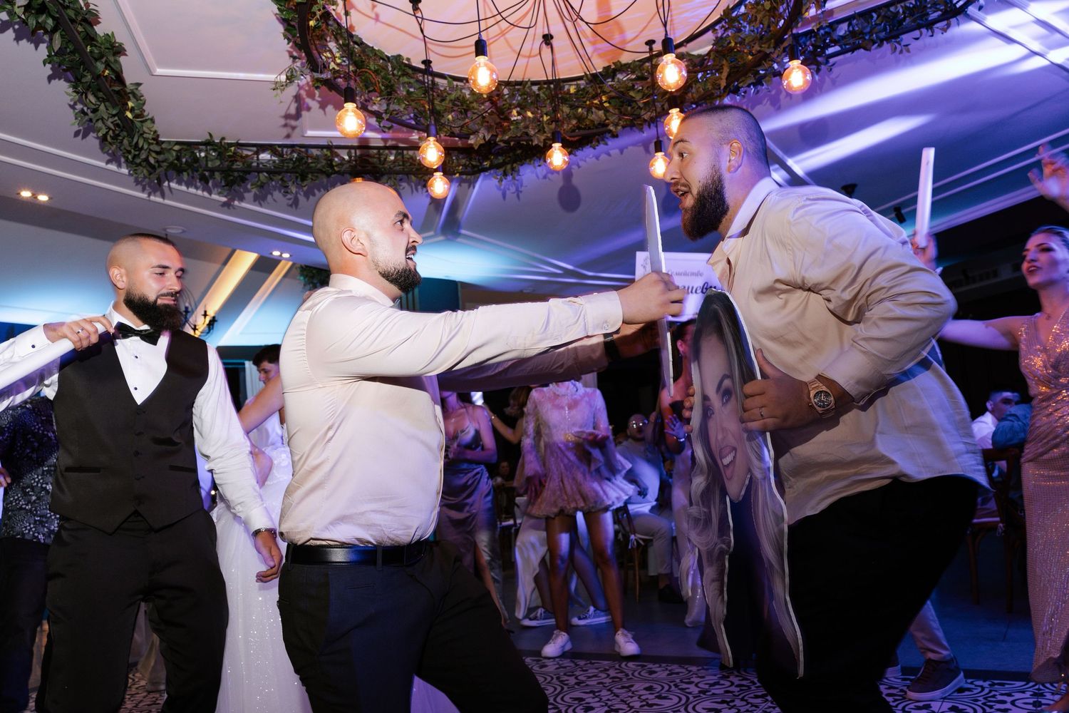 People dancing and celebrating on a dance floor at a wedding reception with colorful purple lighting and decor.