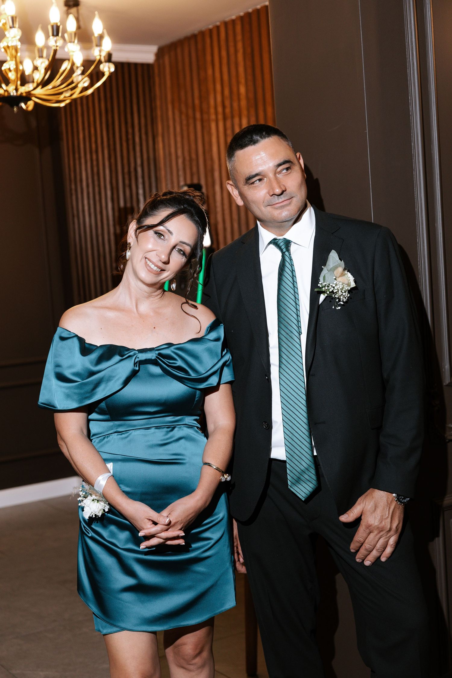A couple in formal attire poses together at an event, with the woman wearing an elegant teal satin dress.