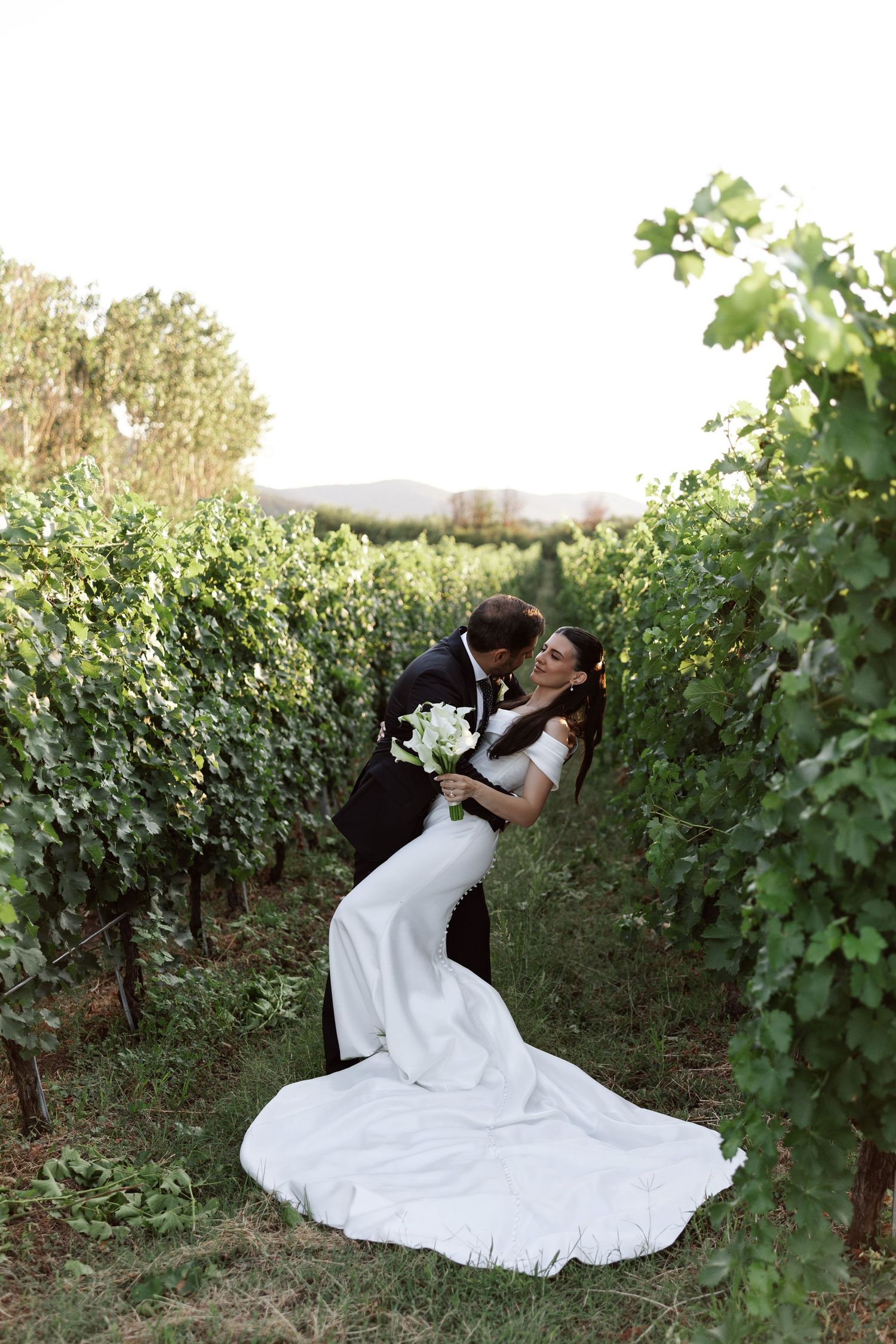 A romantic couple embraces in a lush vineyard setting with a flowing white gown spread across the ground.