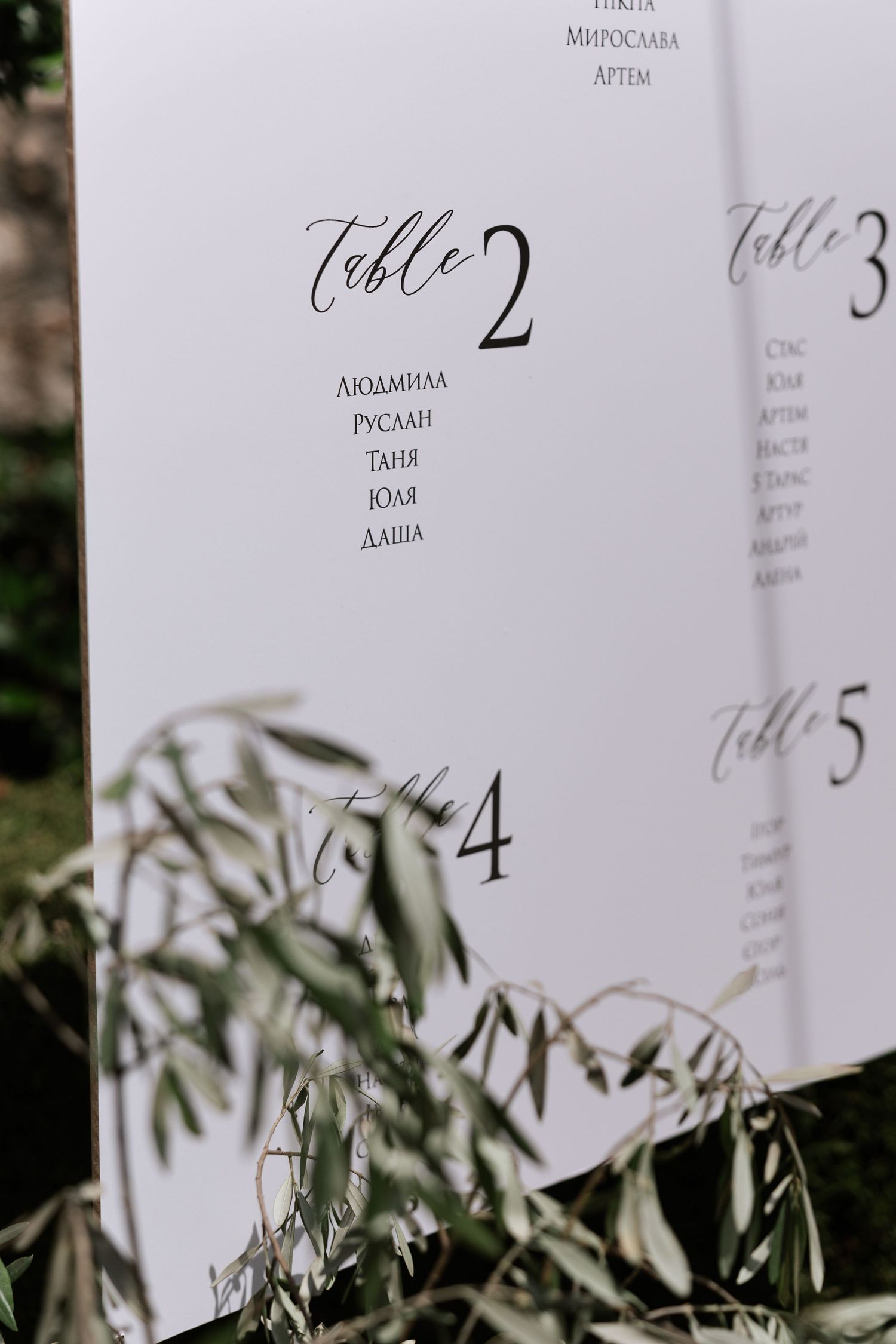 Elegant wedding table number cards with calligraphy displayed among olive branches at a reception.