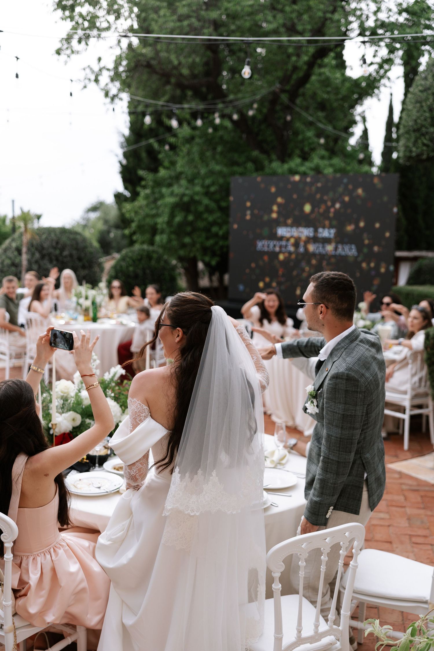 Wedding guests raise their glasses in celebration during an outdoor reception with white chairs and greenery.
