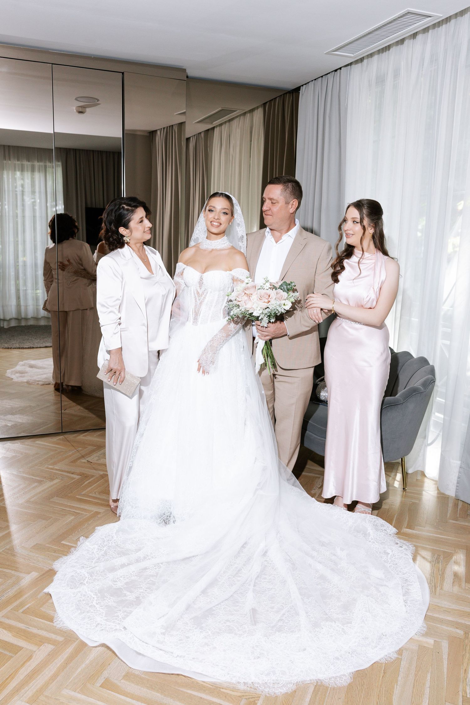 A wedding party in white dresses and formal attire gather in an elegant room with hardwood floors and sheer curtains.