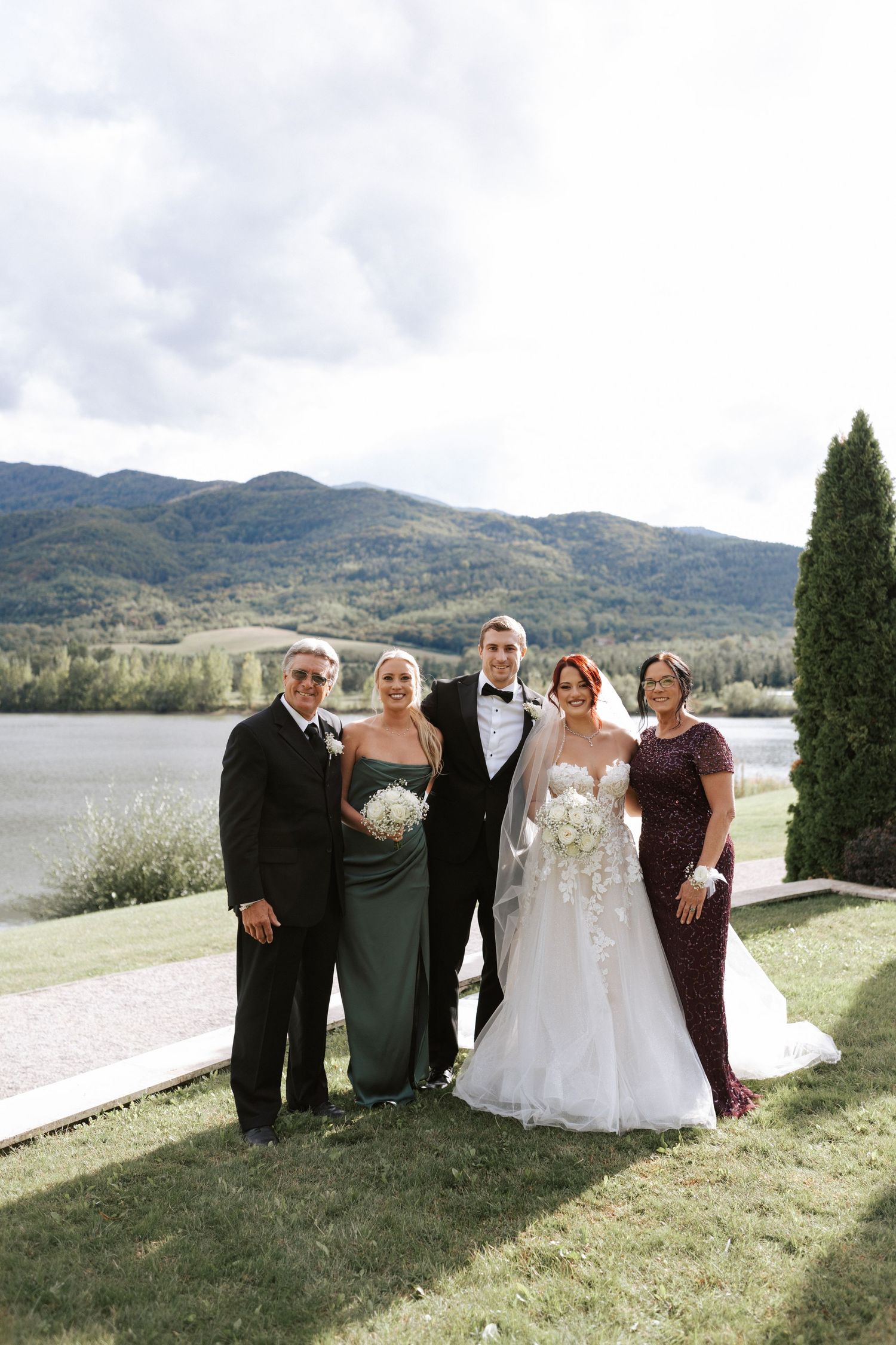 Wedding party group photo on a lawn with mountain and lake backdrop.