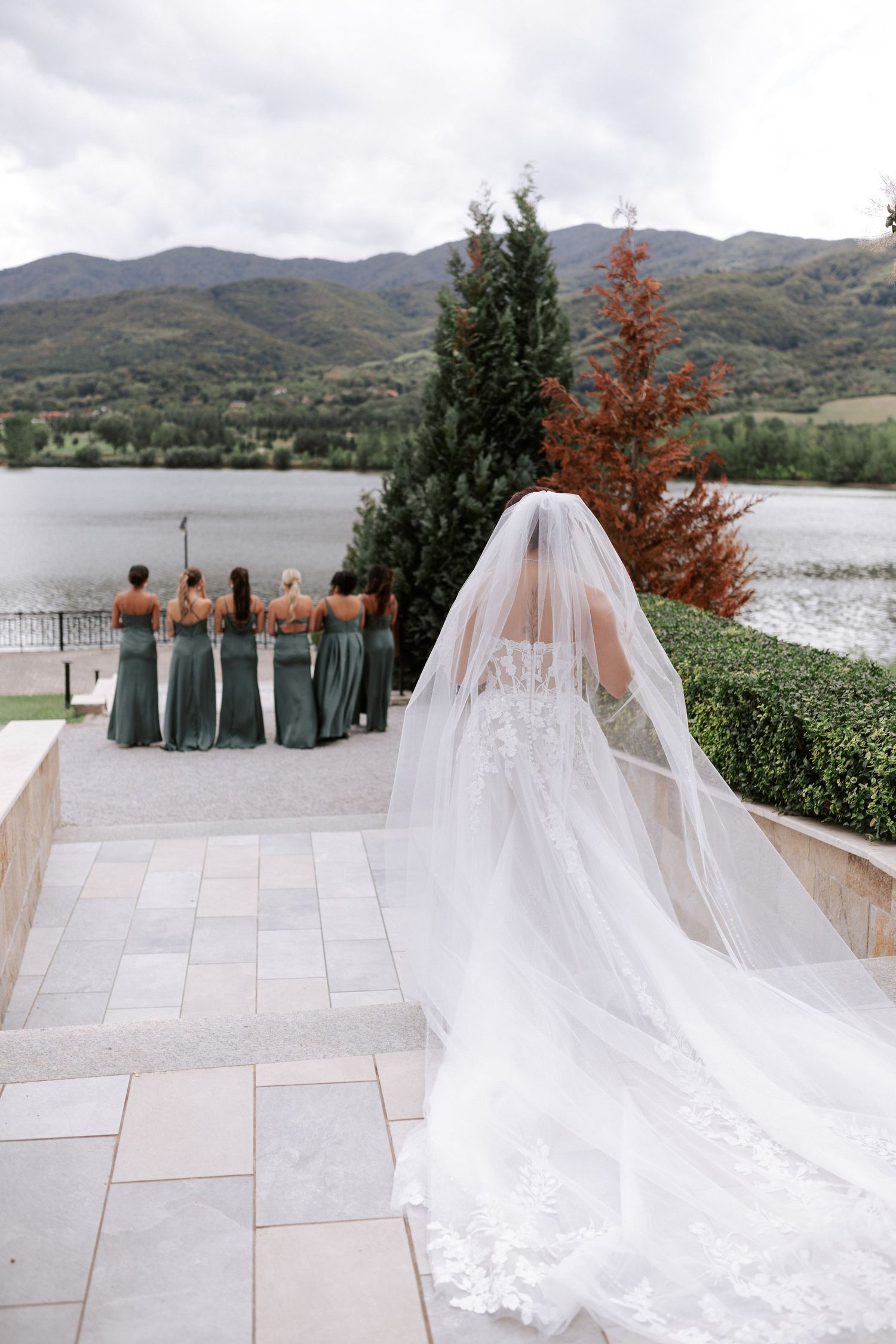 Wedding party photo showing a bride's flowing veil and dress against a mountain lake backdrop.