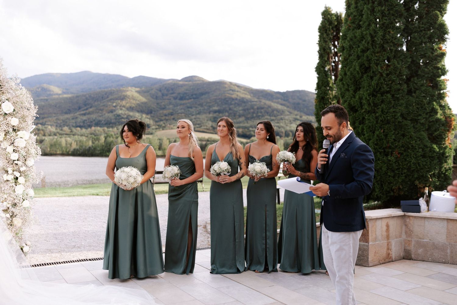 Bridesmaids in elegant sage green dresses stand together on a terrace overlooking mountain scenery.