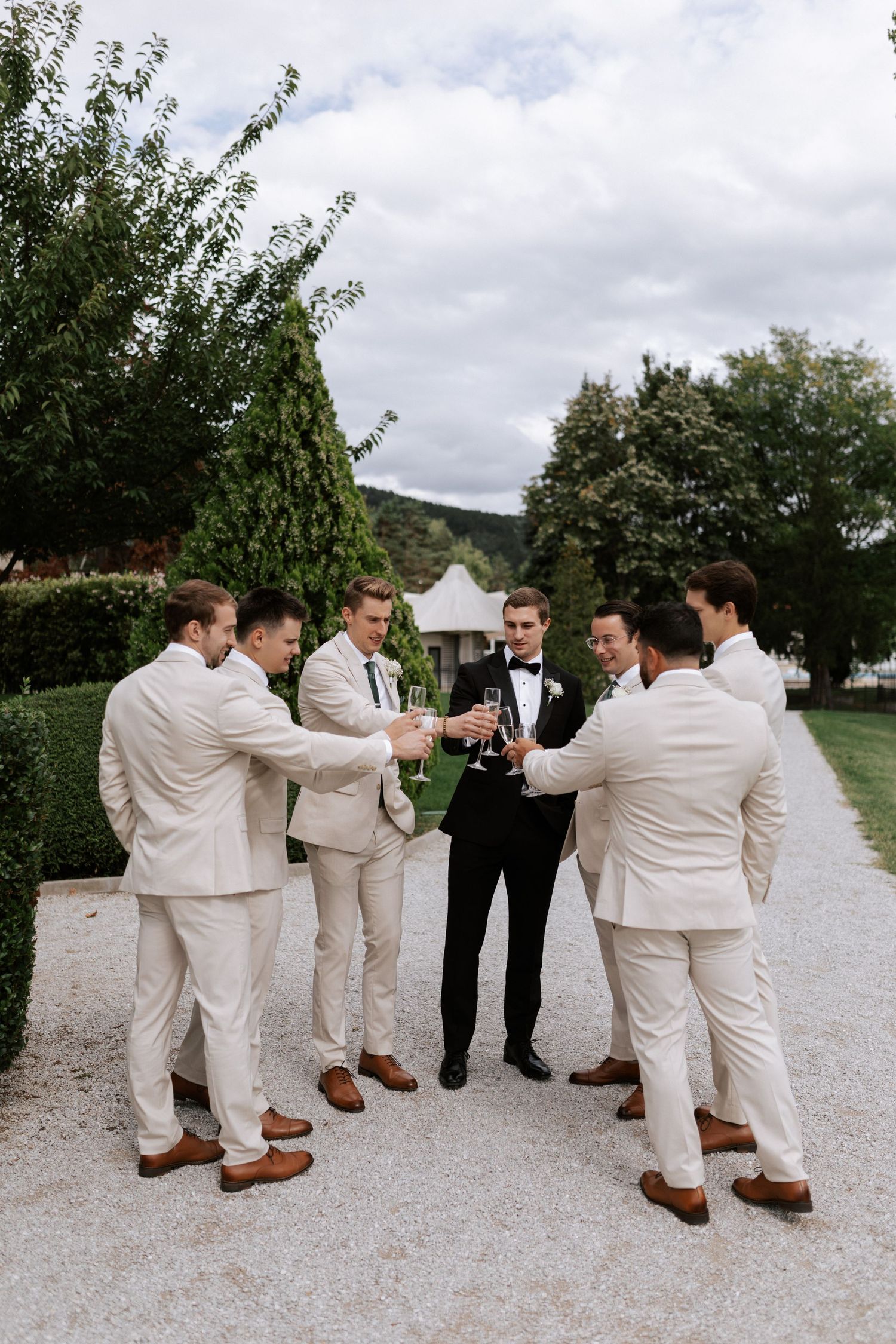 Wedding party in light suits toasts with drinks on garden pathway.