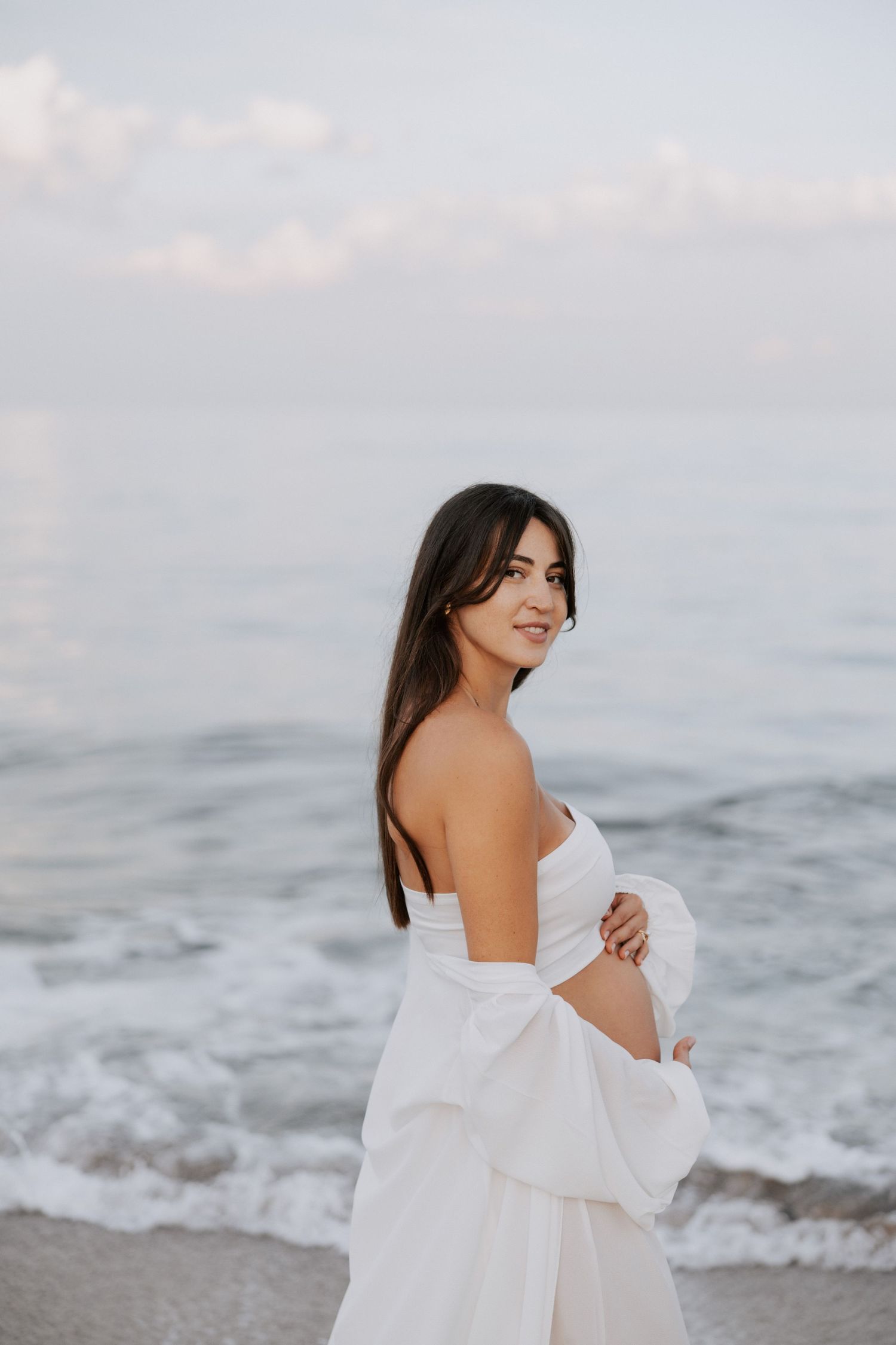 An expectant mother in a white dress poses on the beach during golden hour.